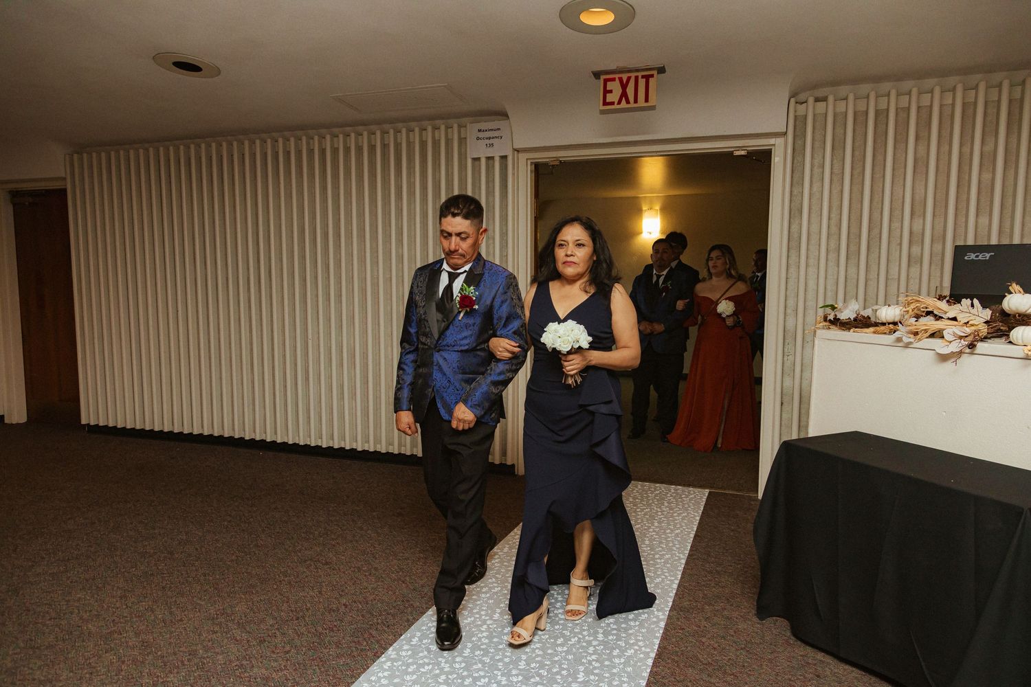 Reception entrance with formal attire couples walking down decorated hallway aisle.
