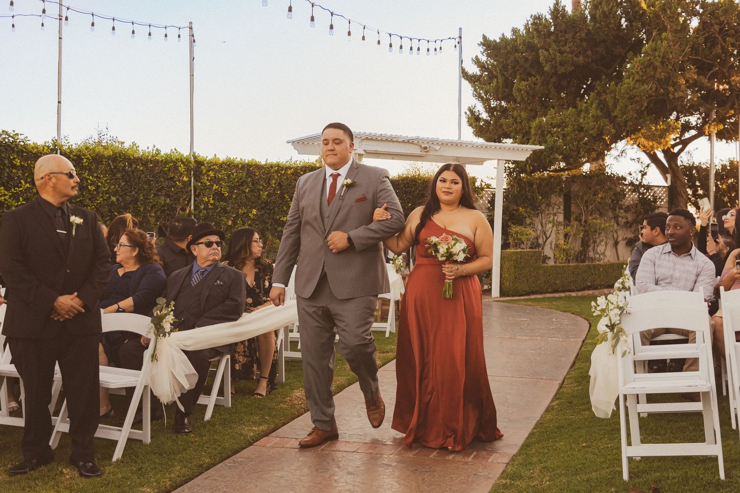 Wedding procession down an aisle decorated with string lights and white folding chairs.