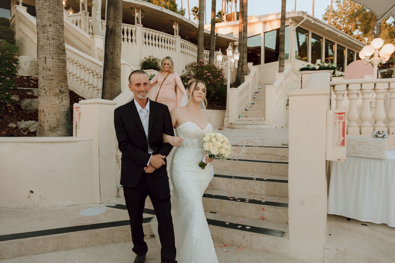 A couple during their wedding ceremony on elegant white stairs with palm trees and string lights overhead.