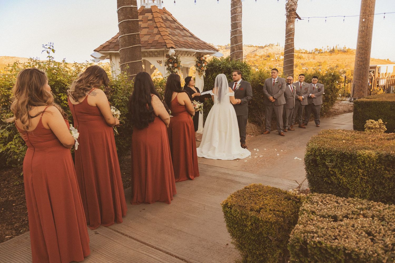 Wedding party gathers at a gazebo with bridesmaids in burgundy dresses and palm trees in the golden sunset.