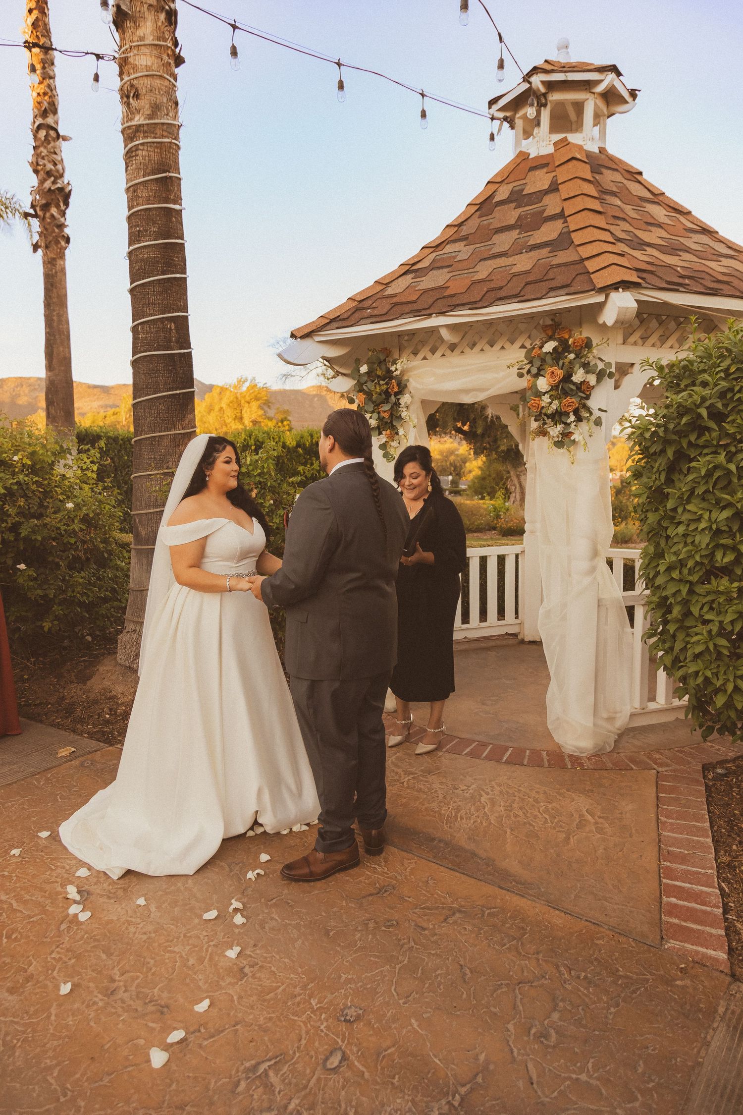 Sunset wedding ceremony taking place in a charming garden gazebo decorated with string lights.
