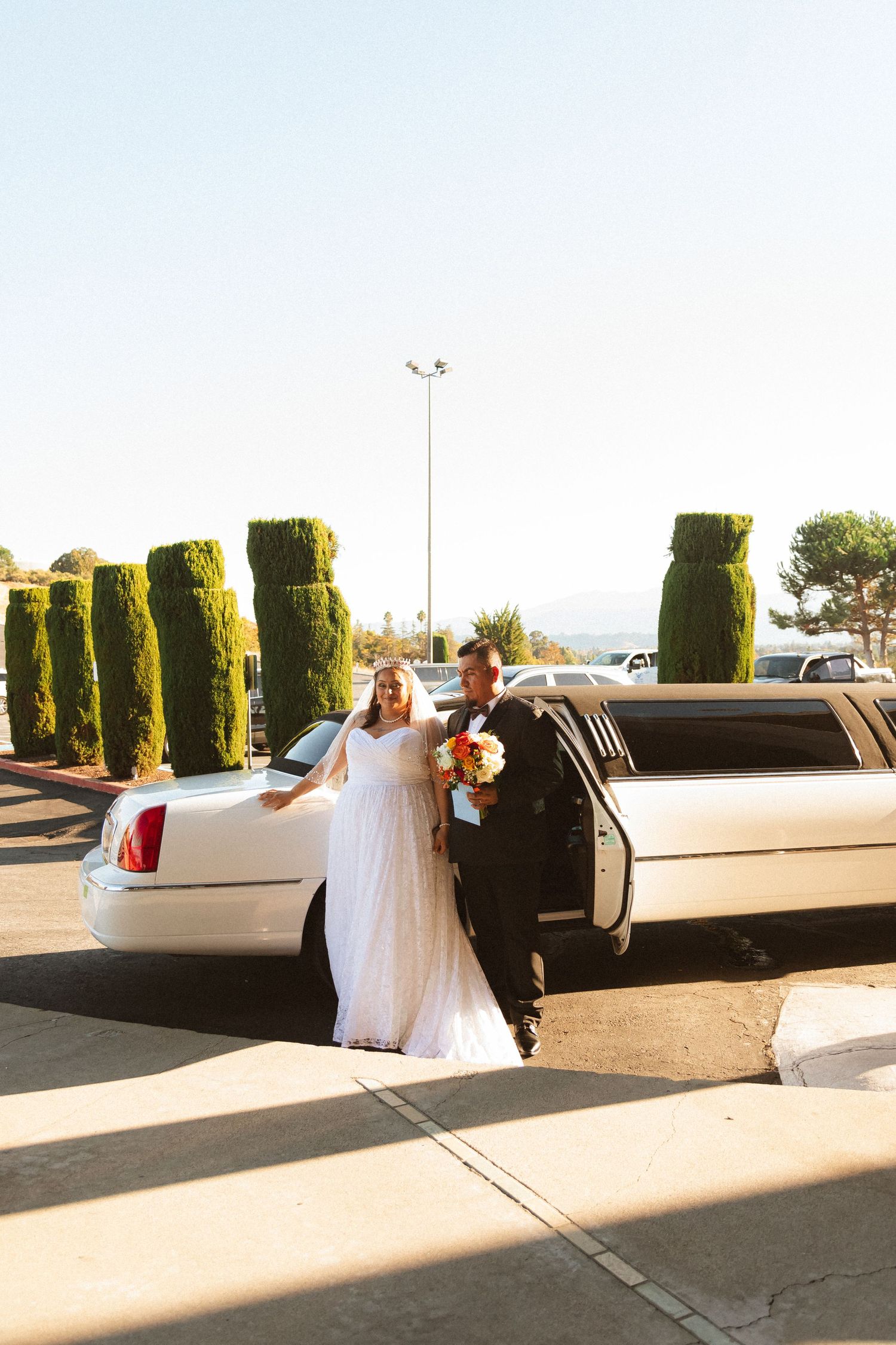 A couple poses by their white limousine with decorative green shrubs in the background.