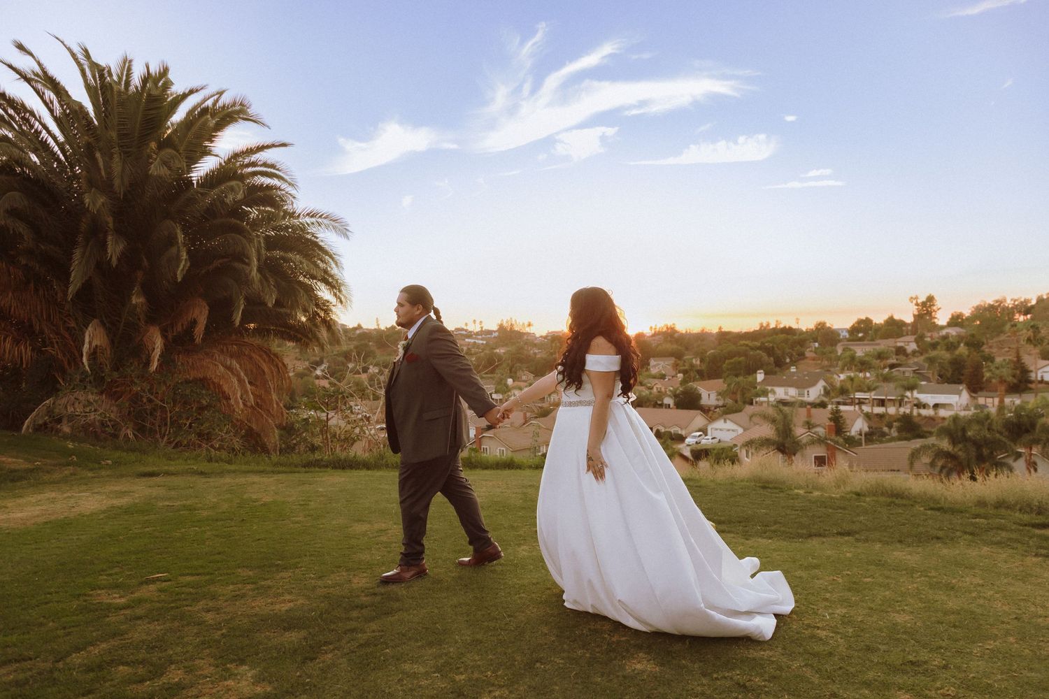 Newlyweds strolling across hilltop lawn with palm trees and cityscape in background.