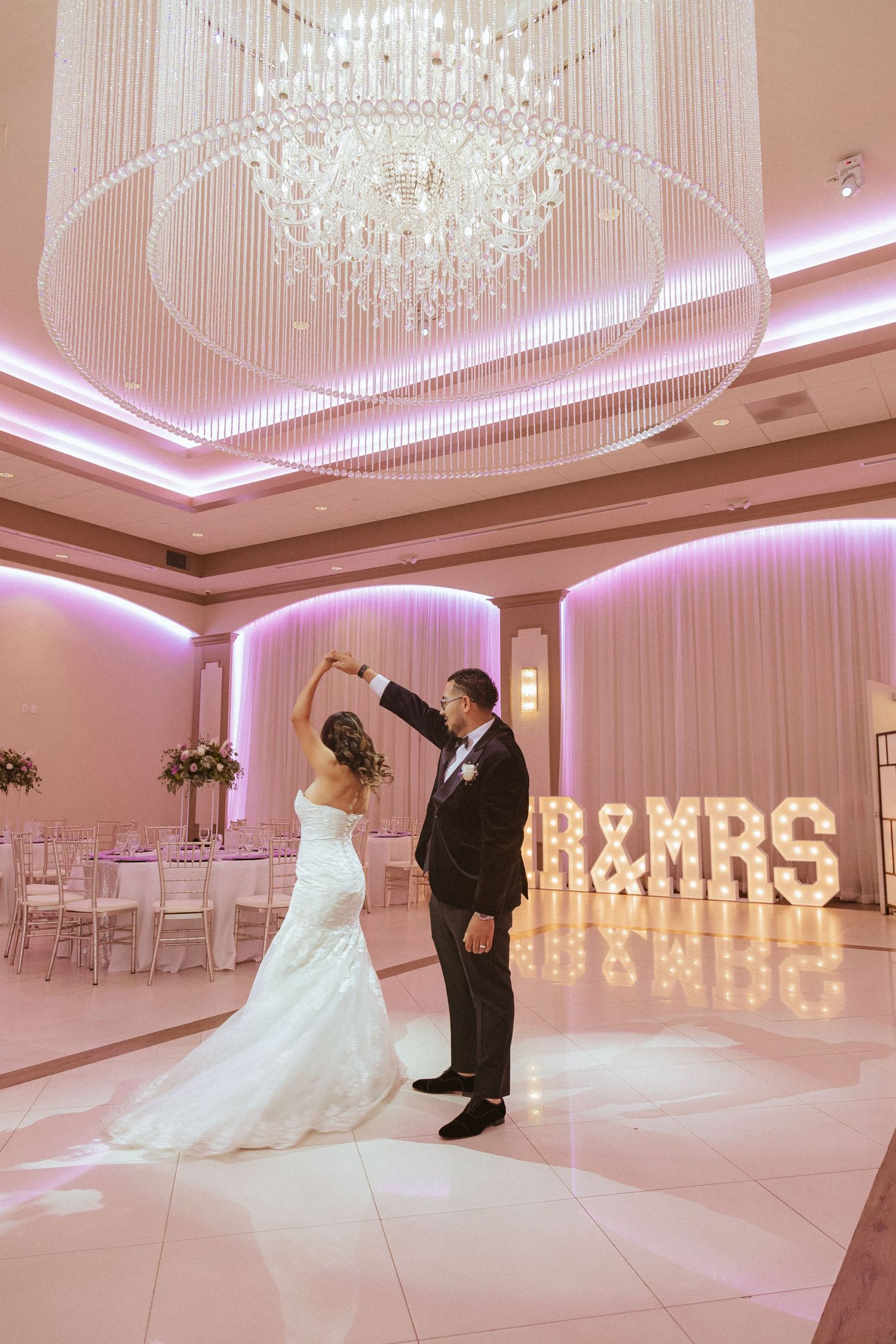 Romantic dance moment in pink-lit ballroom with dramatic chandelier and MR&MRS sign.