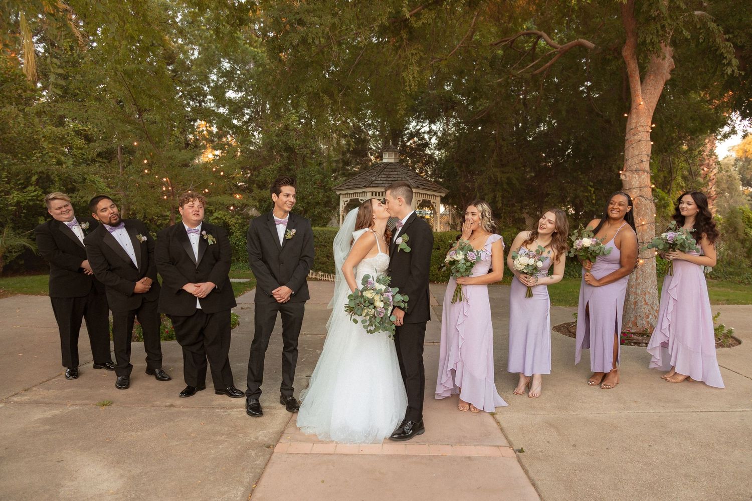 Elegant wedding party poses on stone pathway surrounded by lush green trees.