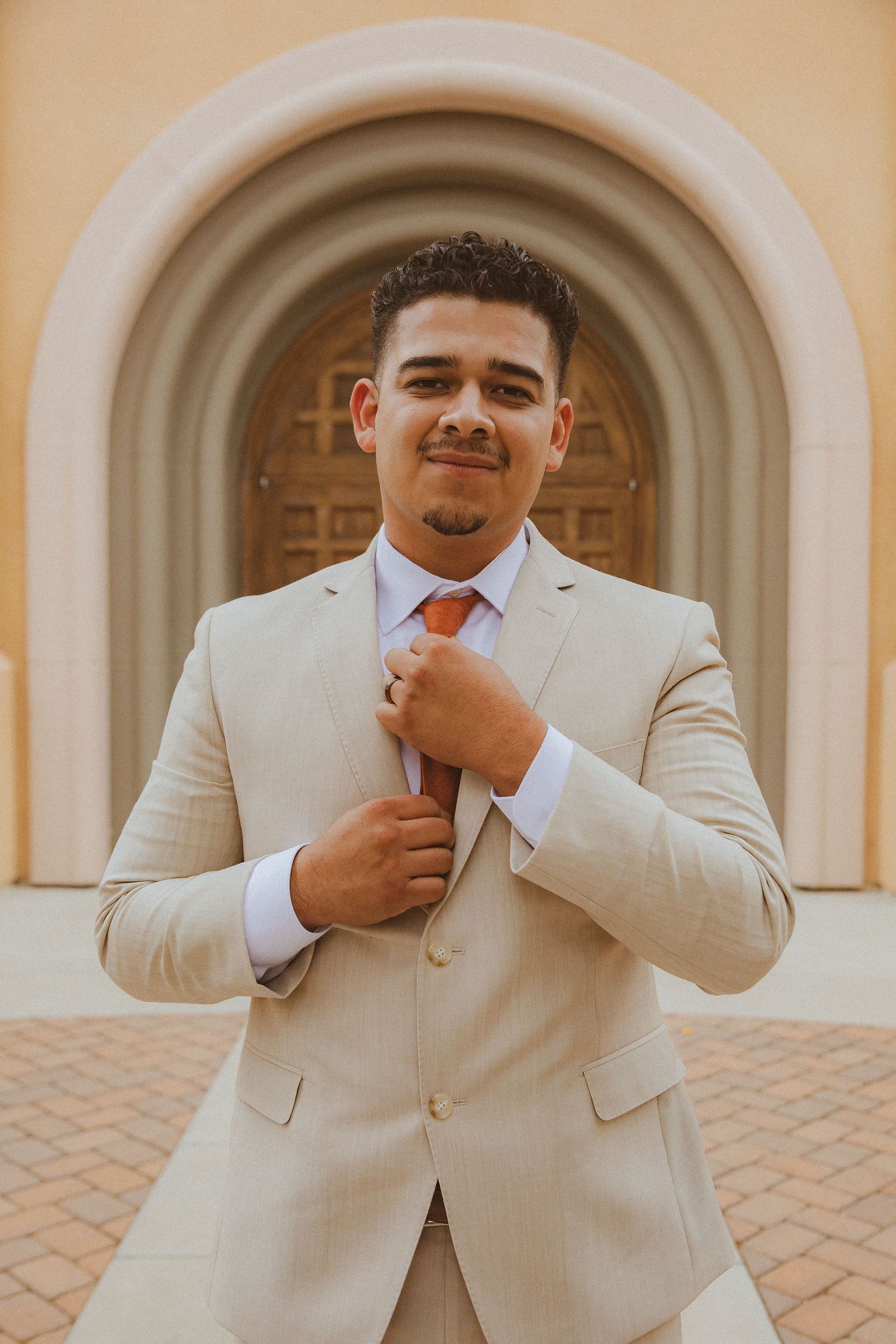 Formal wedding portrait of person in light beige suit adjusting orange tie against arched doorway.