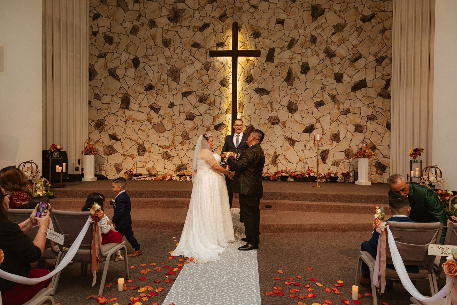 Wedding ceremony in progress at stone-walled church with illuminated cross and rose petal aisle.