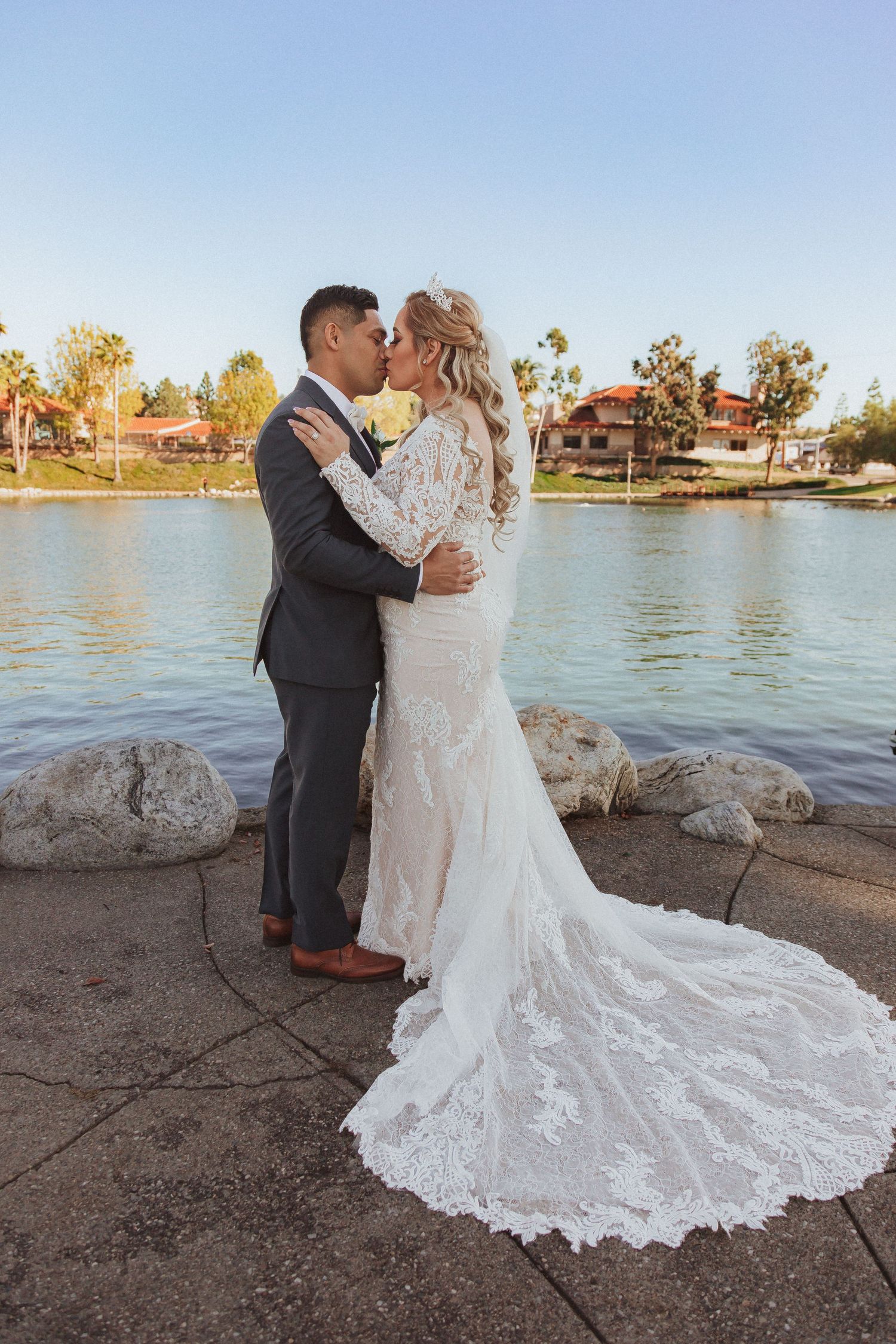 A romantic wedding kiss captured beside a lake with a stunning lace train flowing behind.