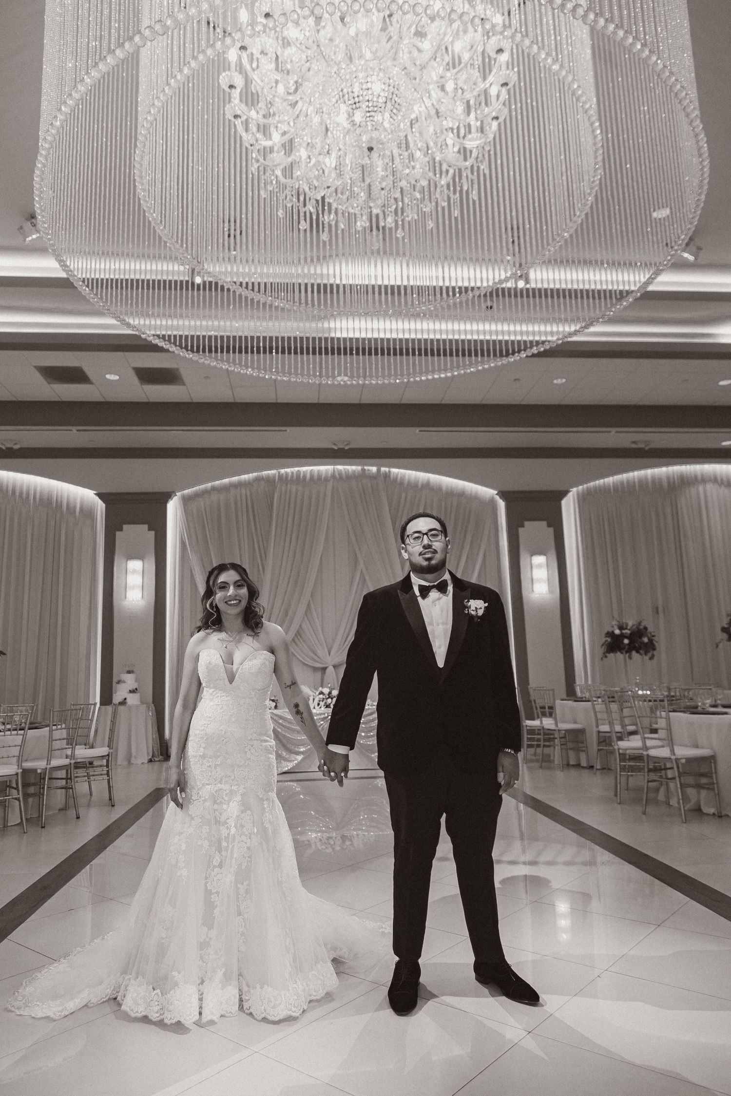 Black and white wedding portrait captures couple holding hands in formal reception setting.