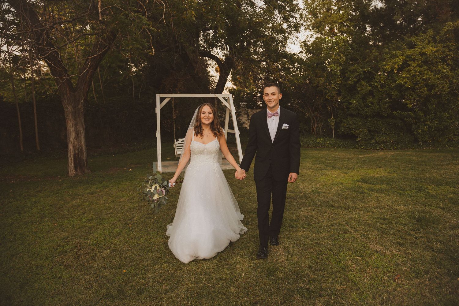 Warm-toned wedding photo of a couple walking together on grass with trees and an arch in the background.