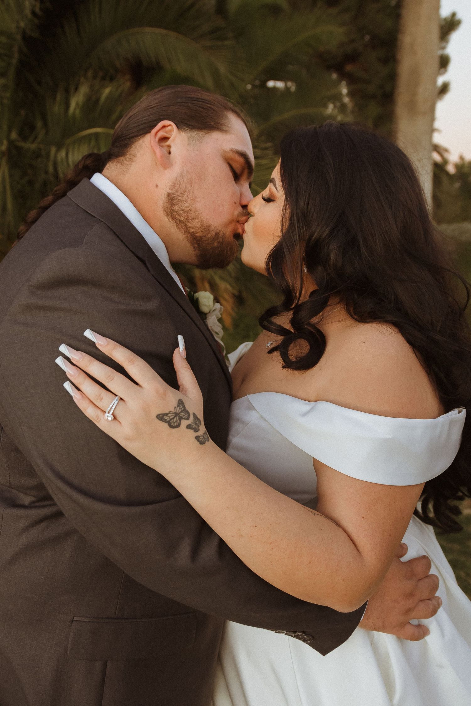 A romantic wedding portrait showing an intimate kiss between newlyweds in a garden setting with palm trees.