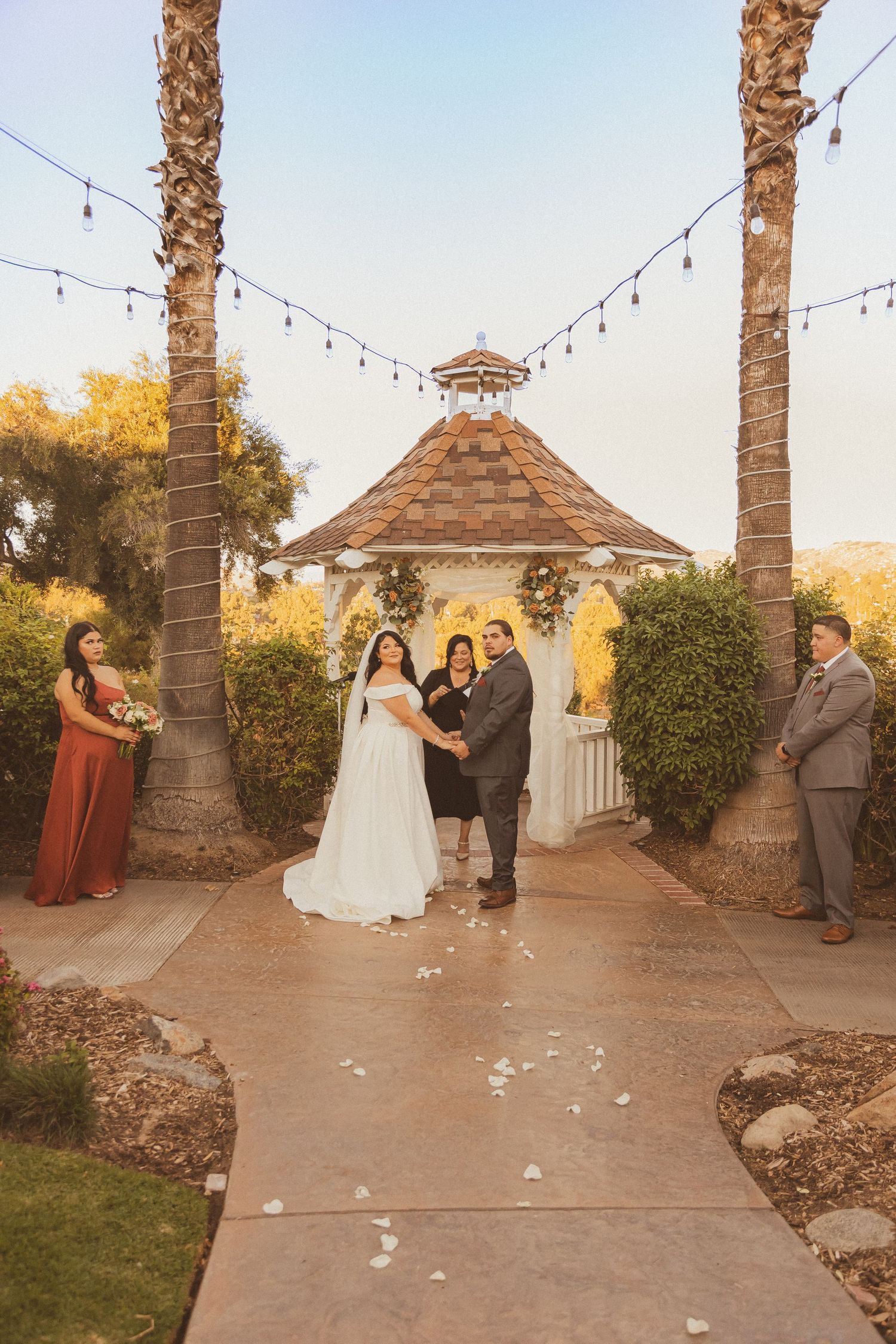 Wedding ceremony in a garden gazebo with palm trees and string lights creating a romantic atmosphere.