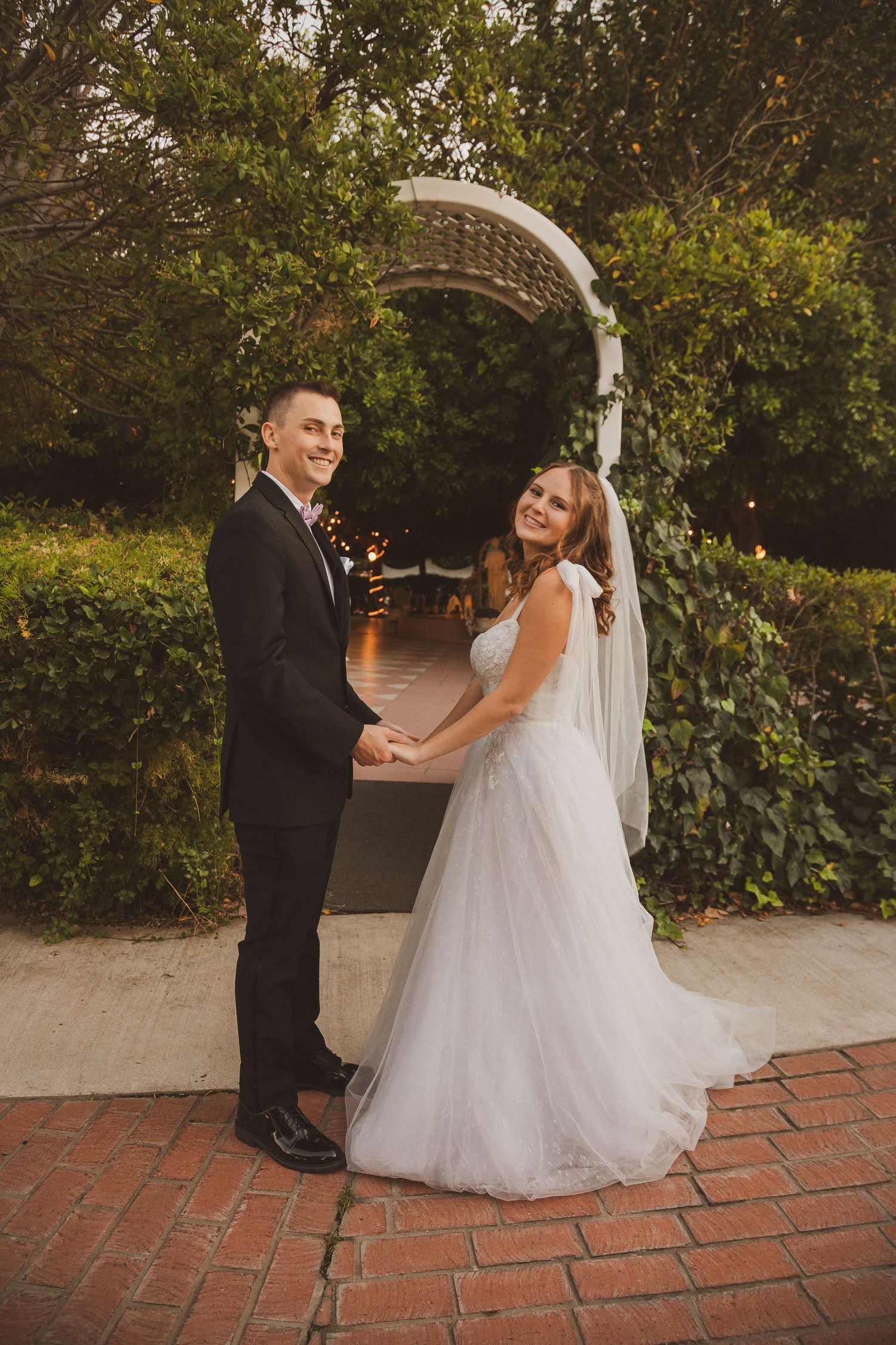 Newlyweds share a romantic moment under an ivy-covered garden arch on brick pavement.