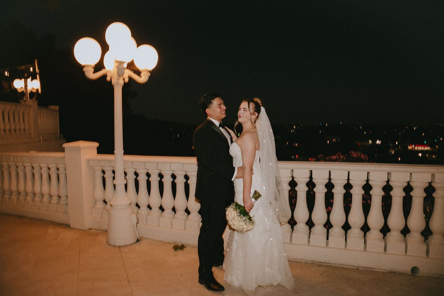 Wedding couple poses against ornate white balustrade with vintage street lamp.