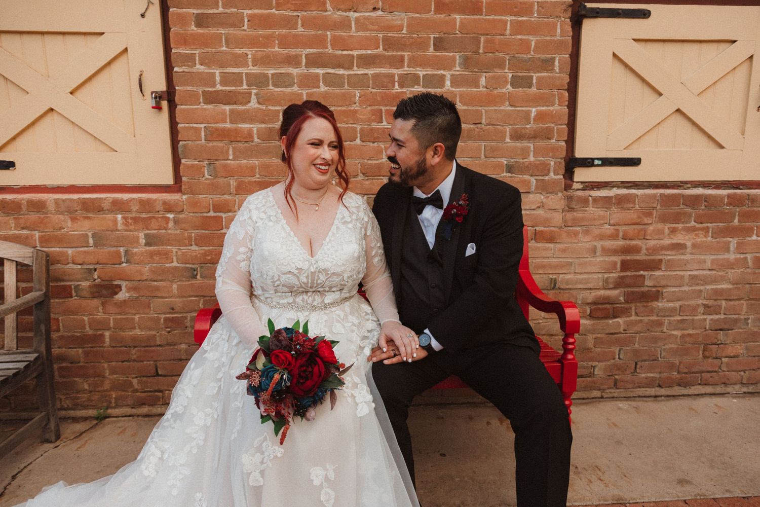 A couple shares a relaxed moment on a wooden bench against a rustic brick wall backdrop.