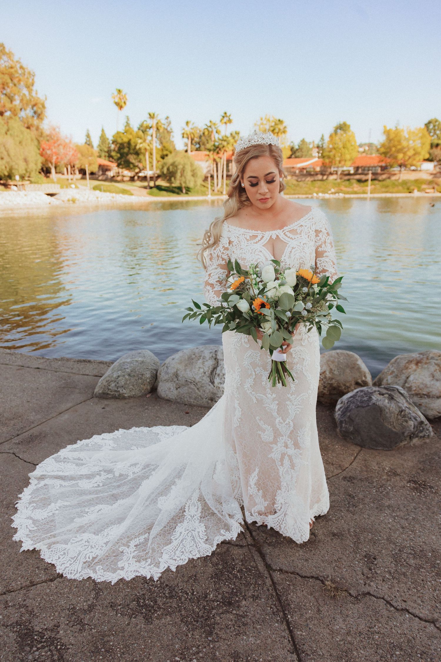Full length bridal photo showing intricate lace wedding dress with long train beside a scenic lakefront.