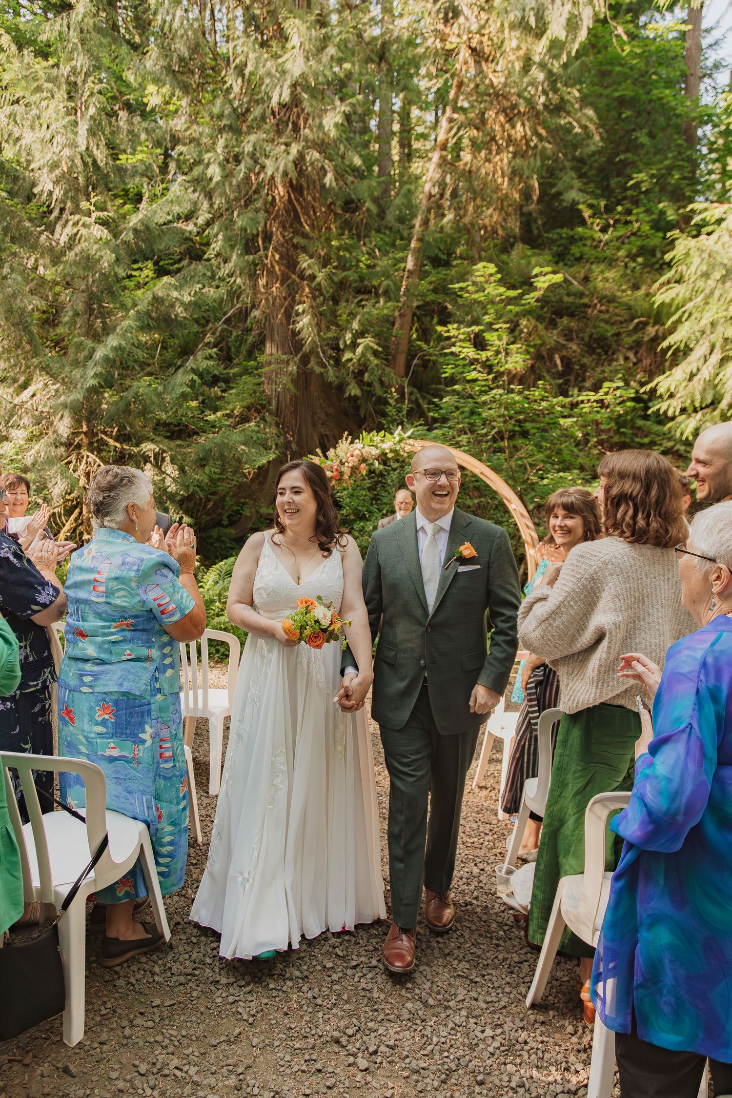 Newlyweds walking down aisle among guests at outdoor forest wedding ceremony.