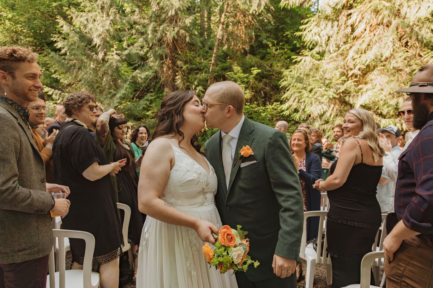 Newlyweds share their first kiss during an outdoor forest wedding ceremony as guests look on.