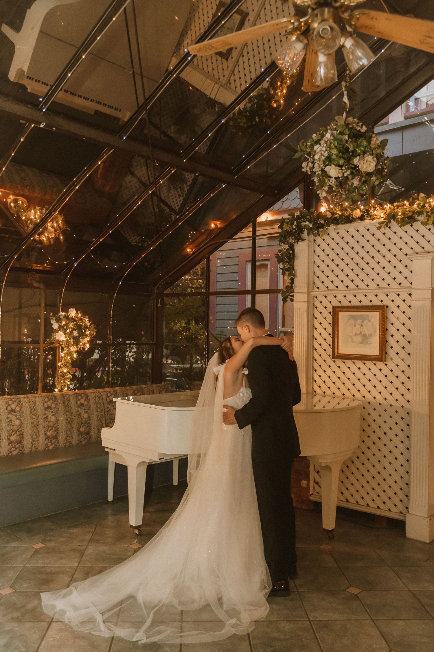 Wedding couple embraces near a white piano under twinkling lights in a glass-ceiling venue.