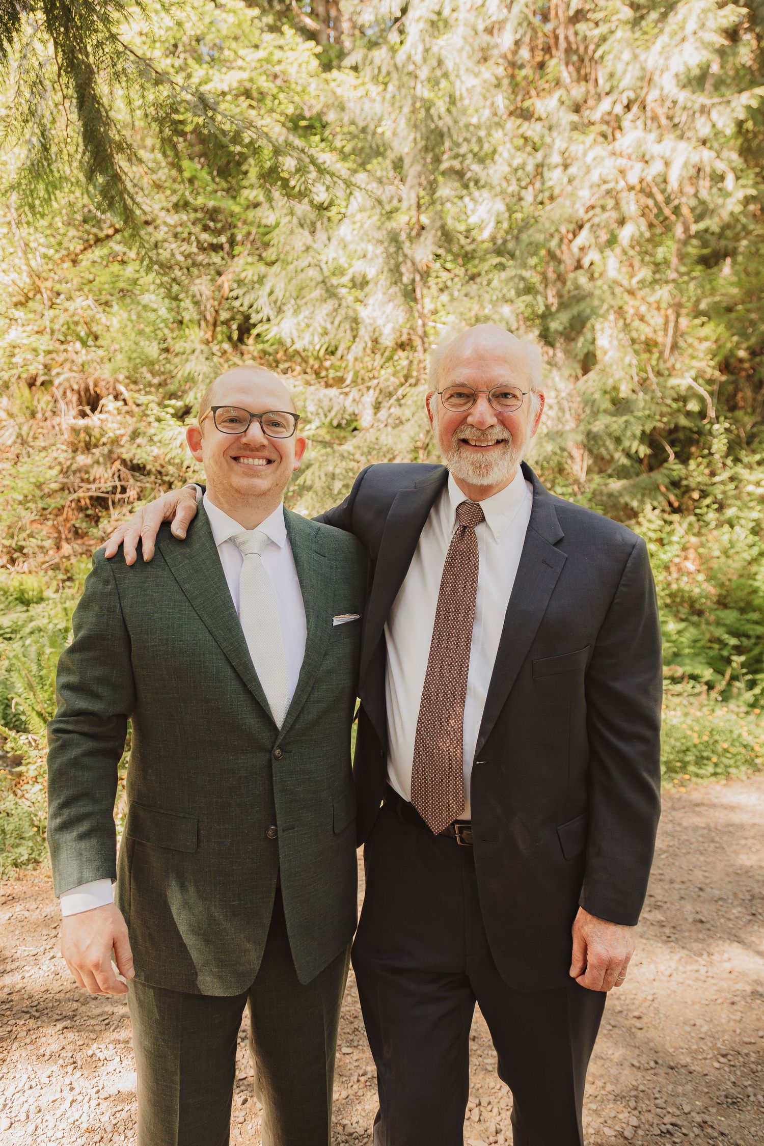 Two men in suits smile warmly for a portrait in a sunlit forest setting.