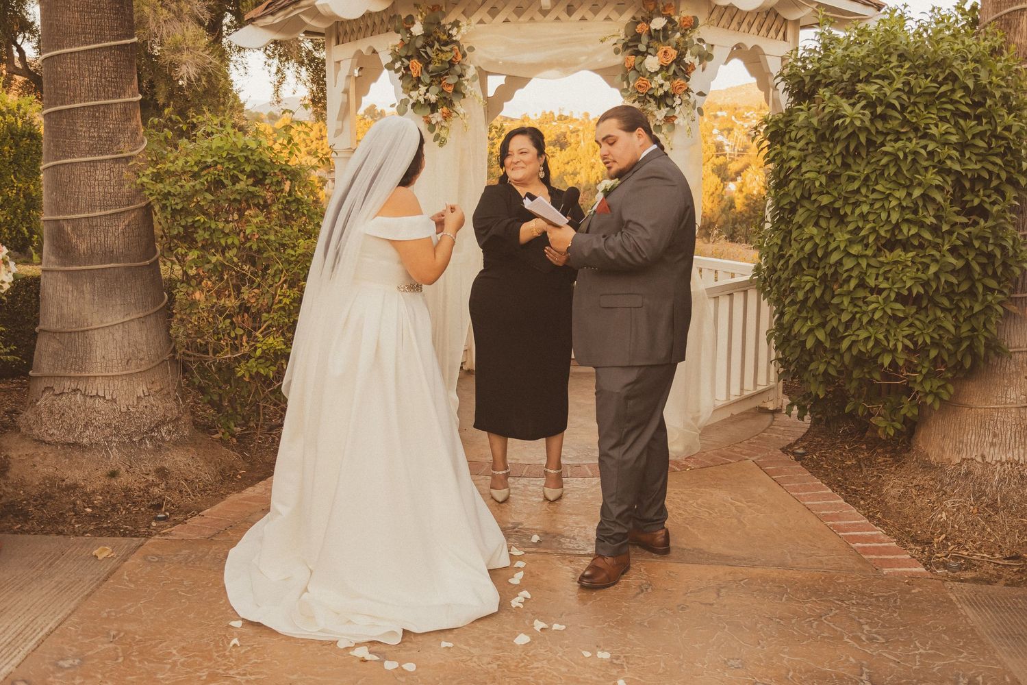 A wedding ceremony takes place in a gazebo with string lights at golden hour.