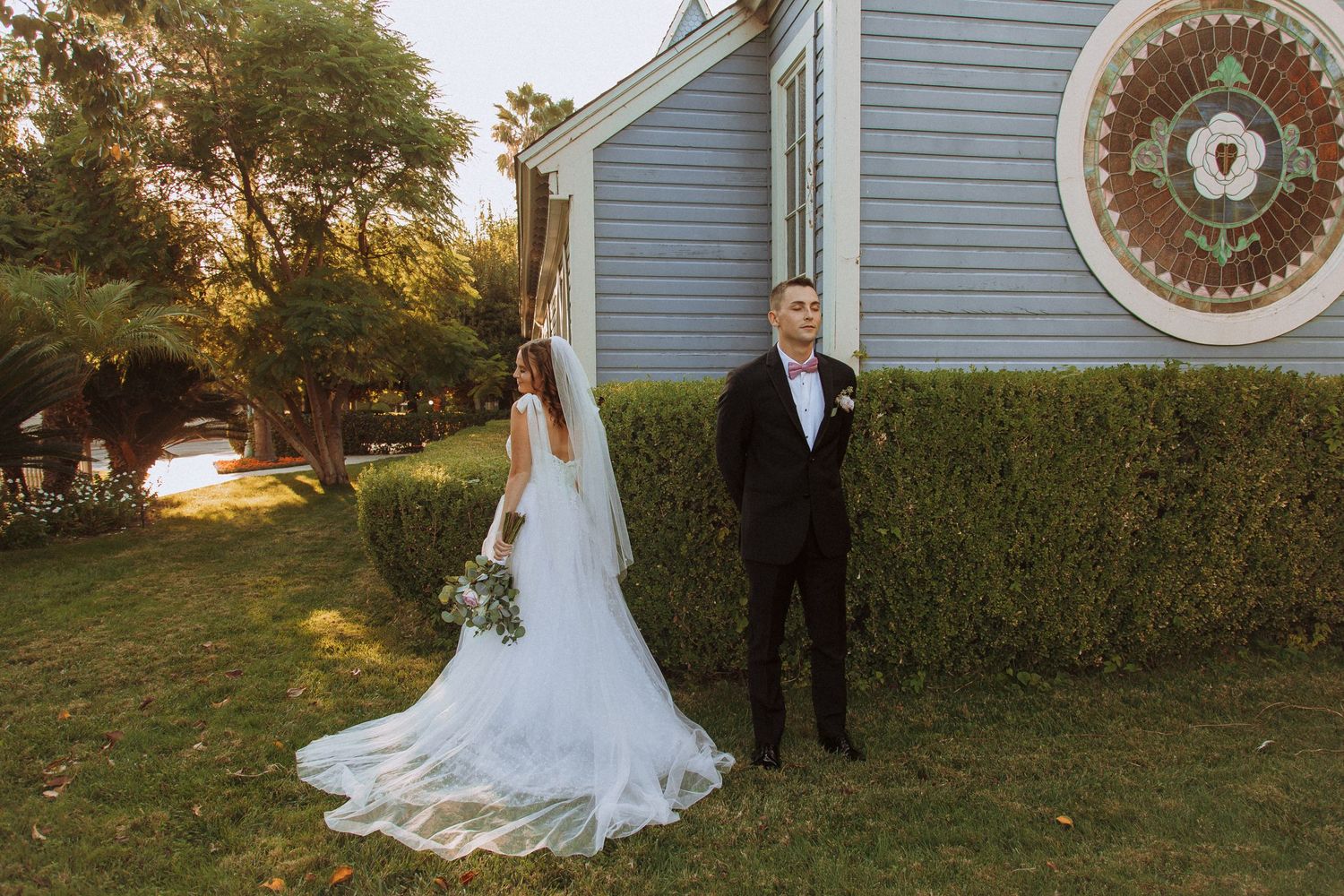 A wedding couple poses in front of a blue church building with hedges at golden hour.