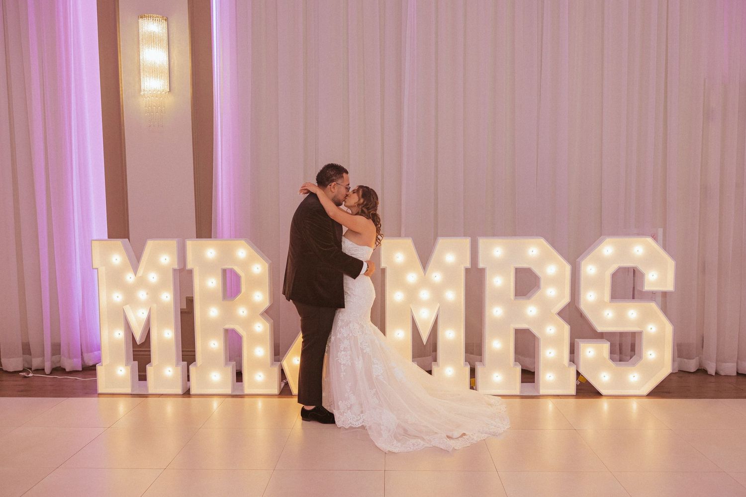 A romantic wedding moment with the couple kissing in front of illuminated MR & MRS marquee letters.