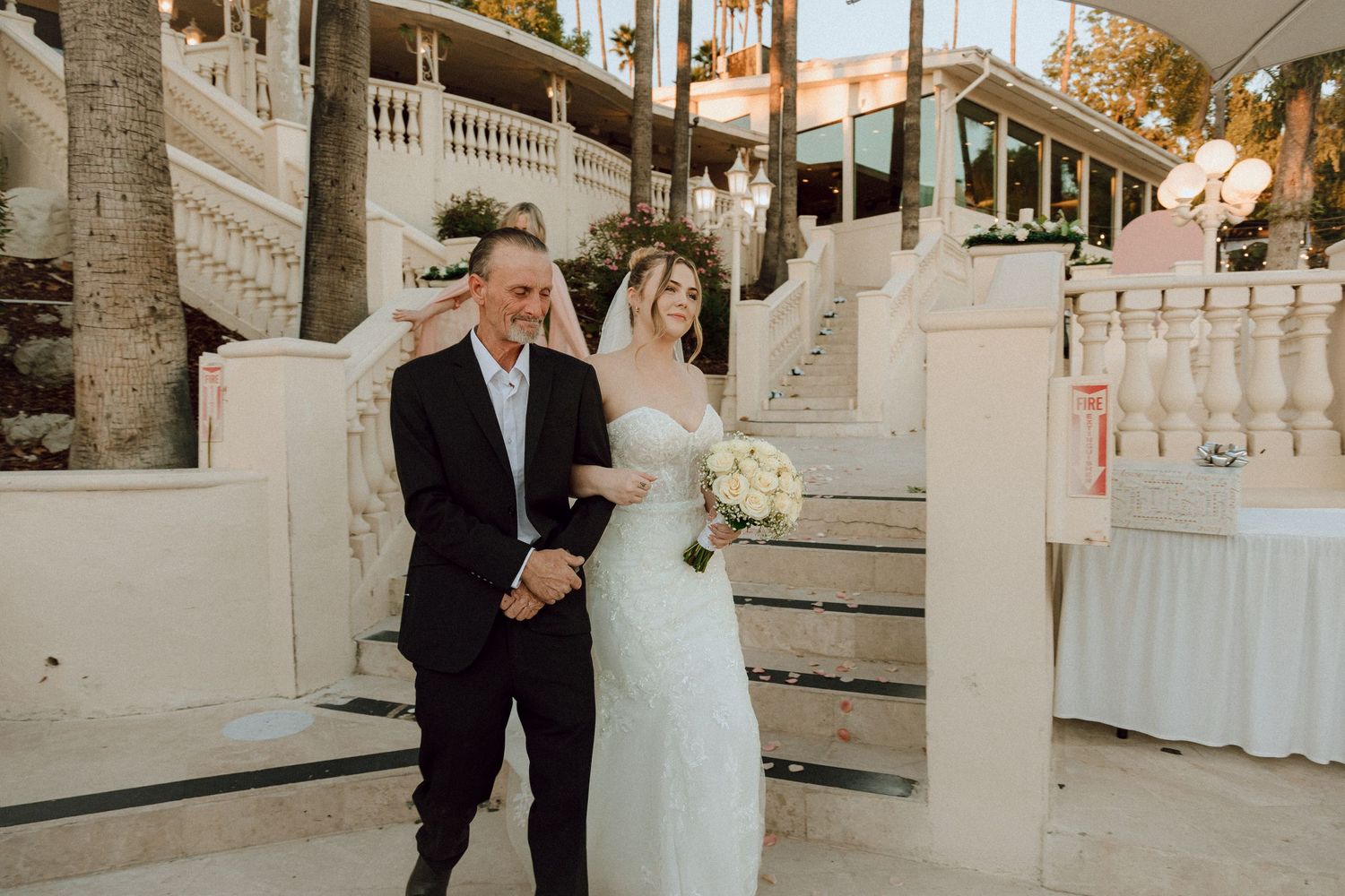 Wedding couple descending ornate white stairs at a tropical venue with warm sunset lighting.