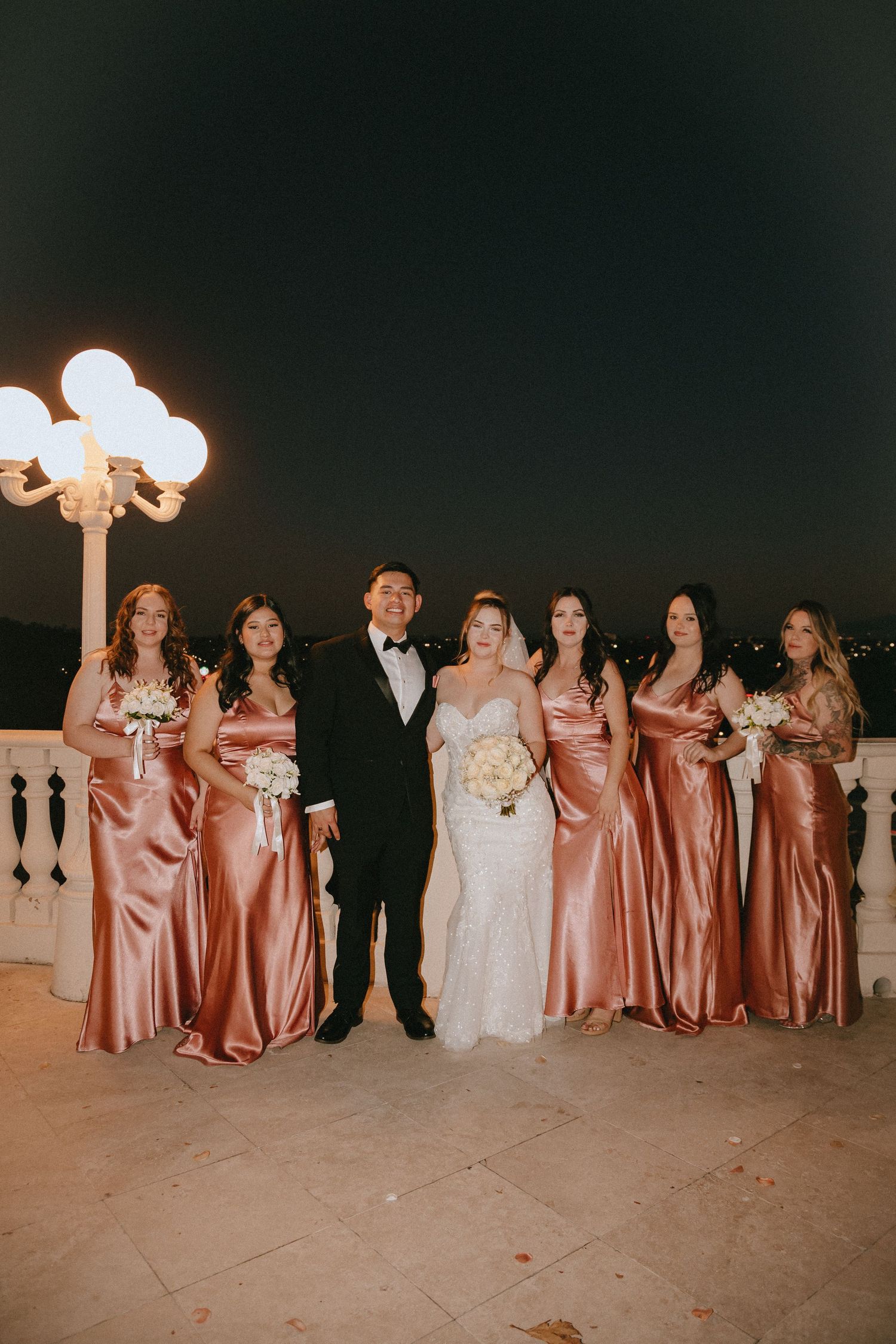 Wedding party poses at night with bridesmaids in rose gold dresses under ornate lighting.