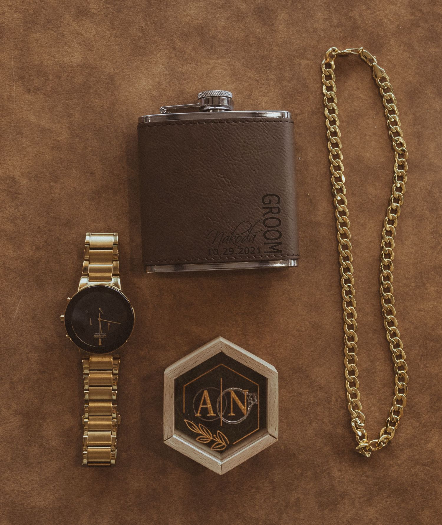 Leather flask, gold watch, monogram logo box and gold chain displayed on brown background.