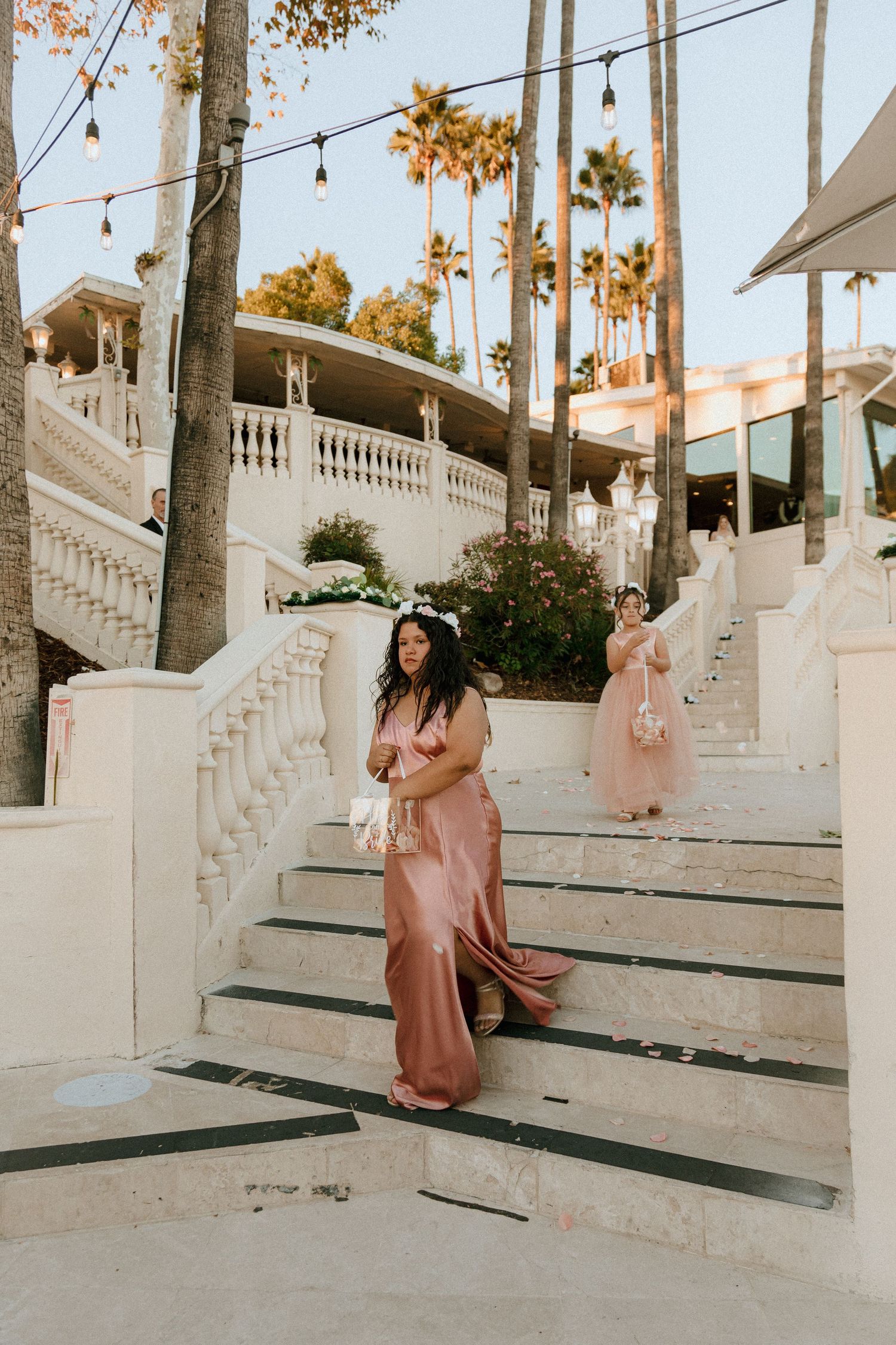 Person in pink satin dress poses on grand white staircase with palm trees and string lights.