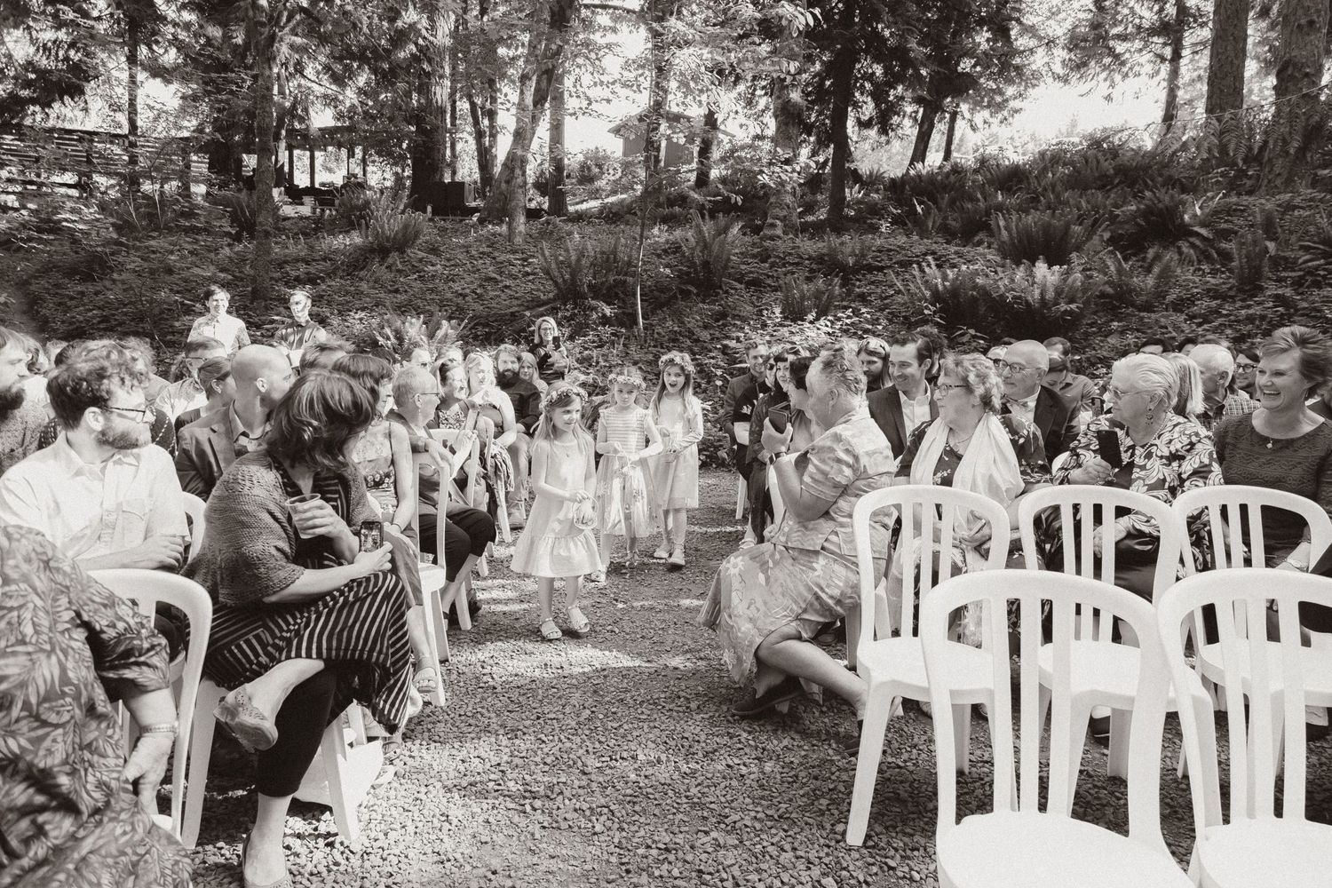 Wedding guests seated in white chairs along a wooded outdoor ceremony path.