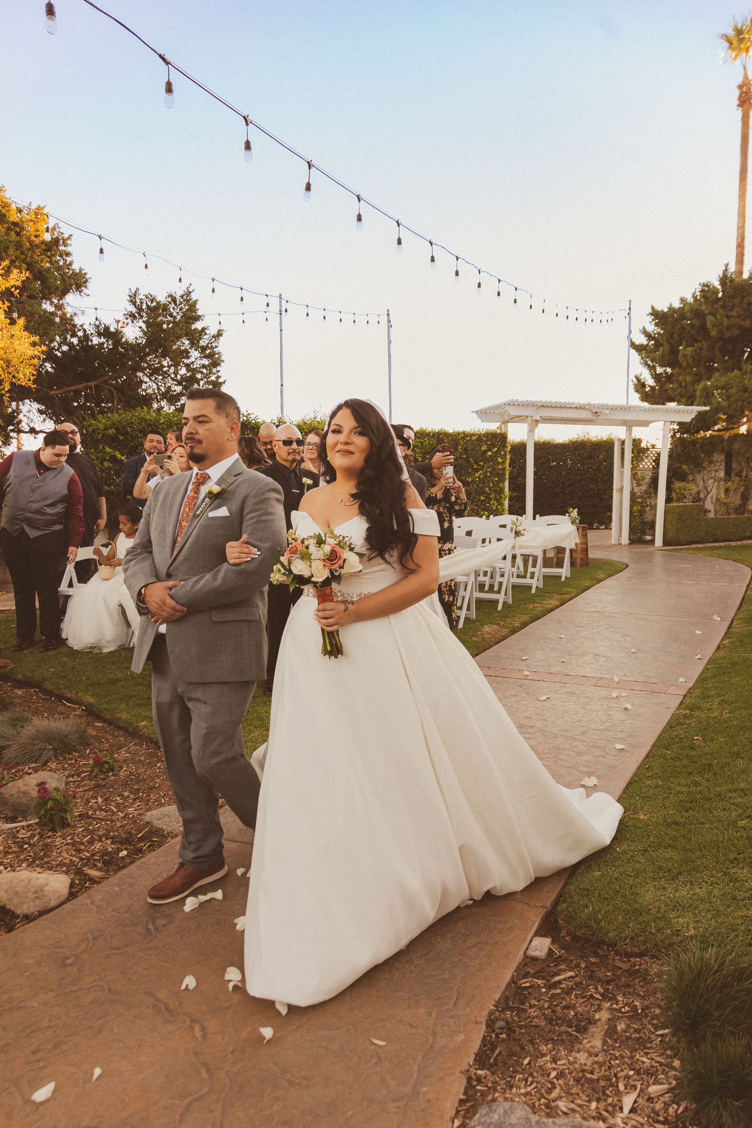Newlyweds walking down garden path under string lights after outdoor ceremony.