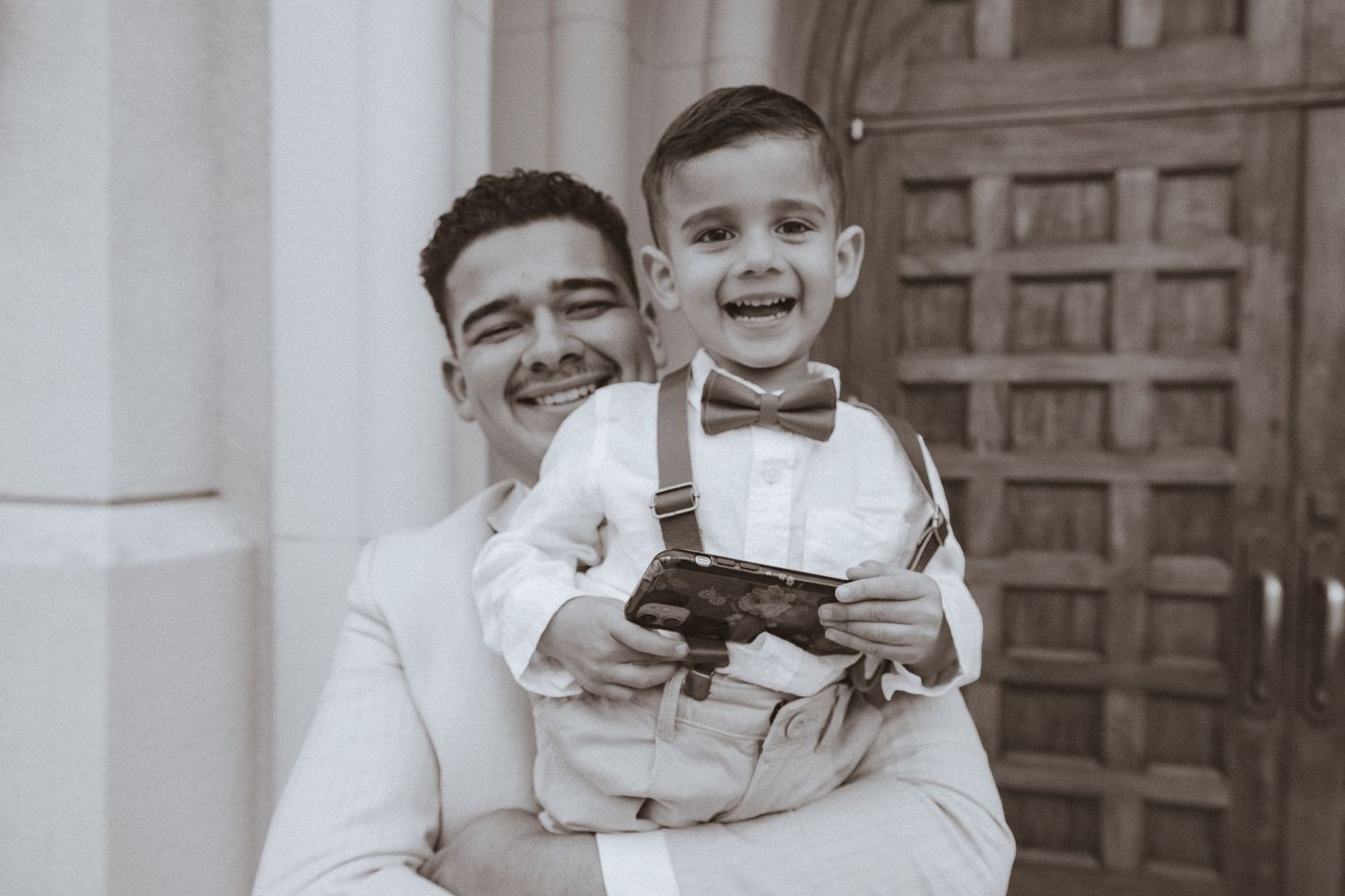 A black and white portrait of a small boy in suspenders and bowtie holding a vintage camera.