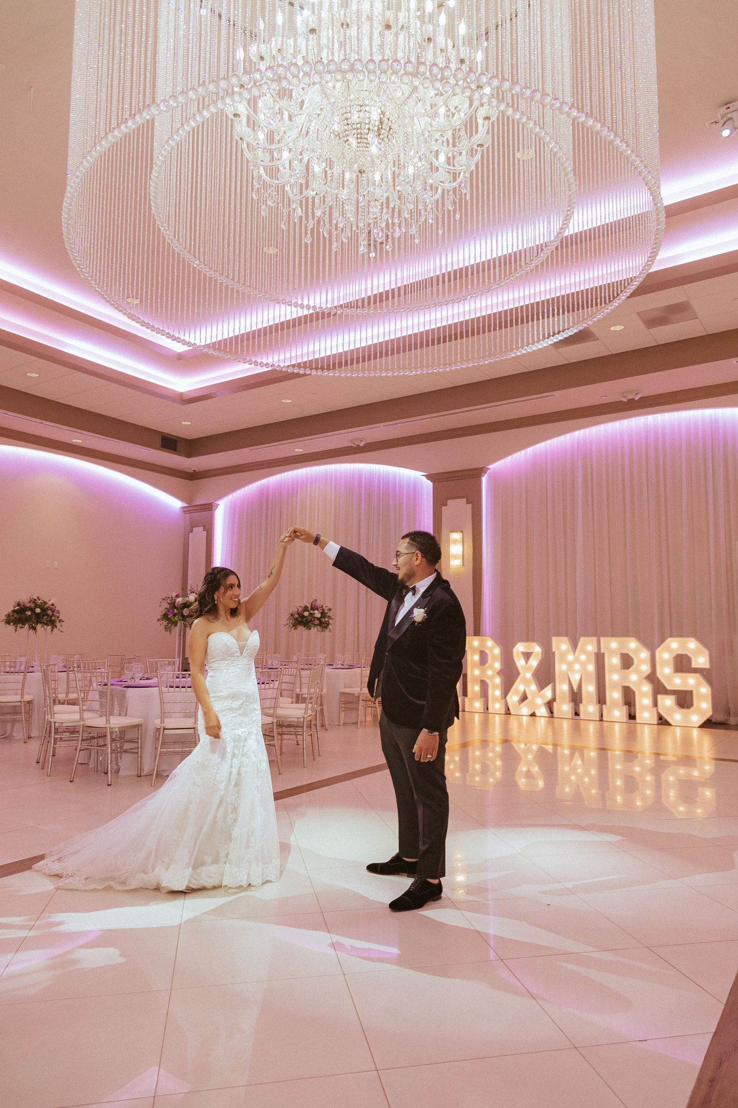 Elegant ballroom wedding scene with couple dancing under crystal chandelier and illuminated signage.