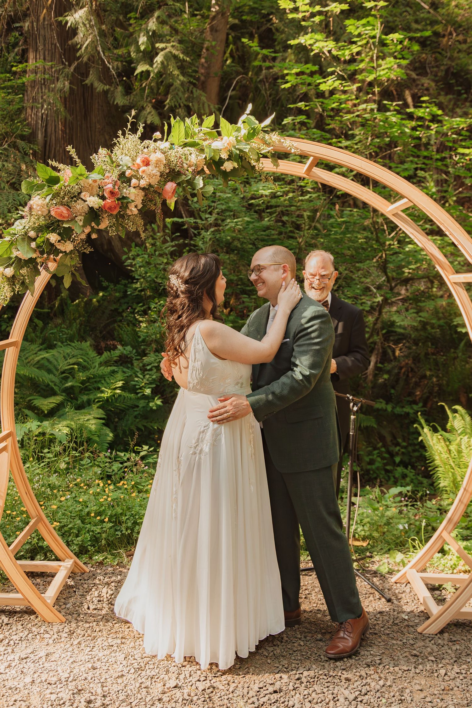 Wedding ceremony beneath wooden circular arch adorned with pink and white flowers in forest setting.