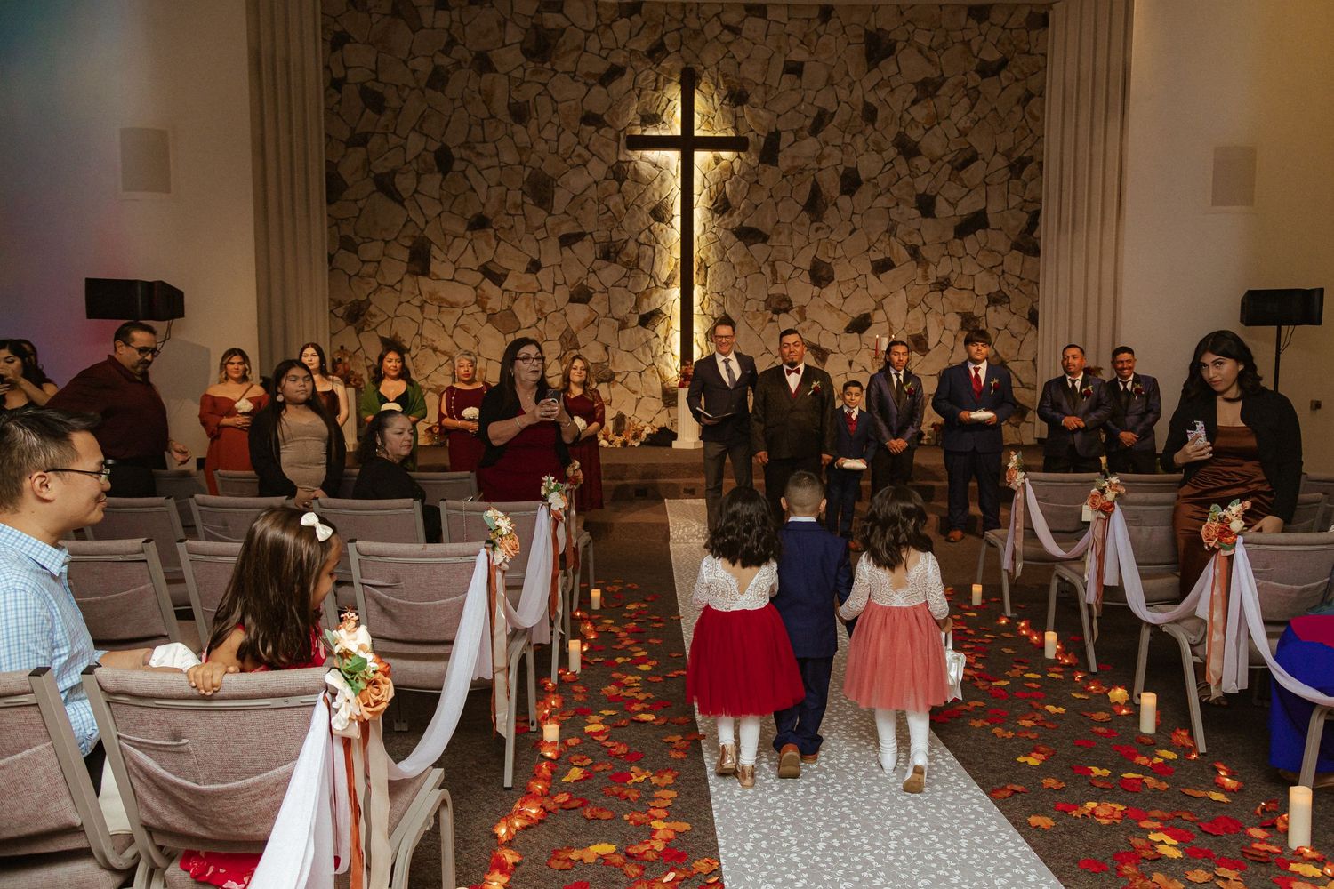 Church wedding ceremony with illuminated cross on floral wallpaper and decorated pews.
