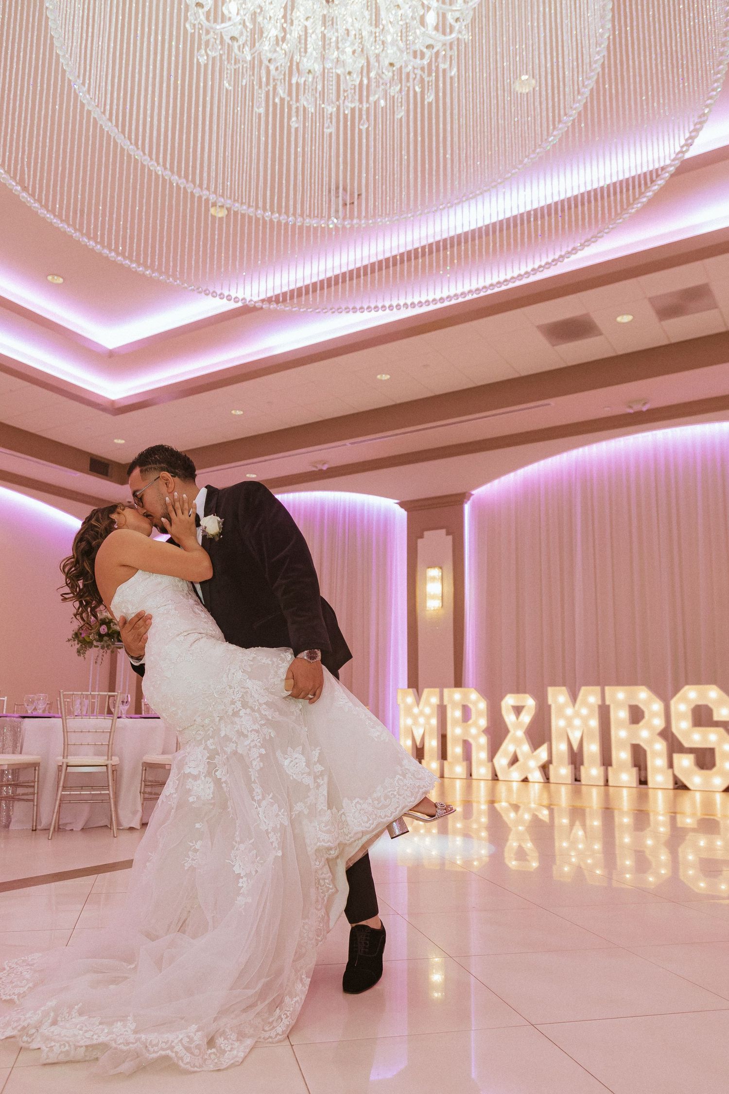 Romantic wedding dip pose captured in elegant ballroom with pink ambient lighting and lit signage.