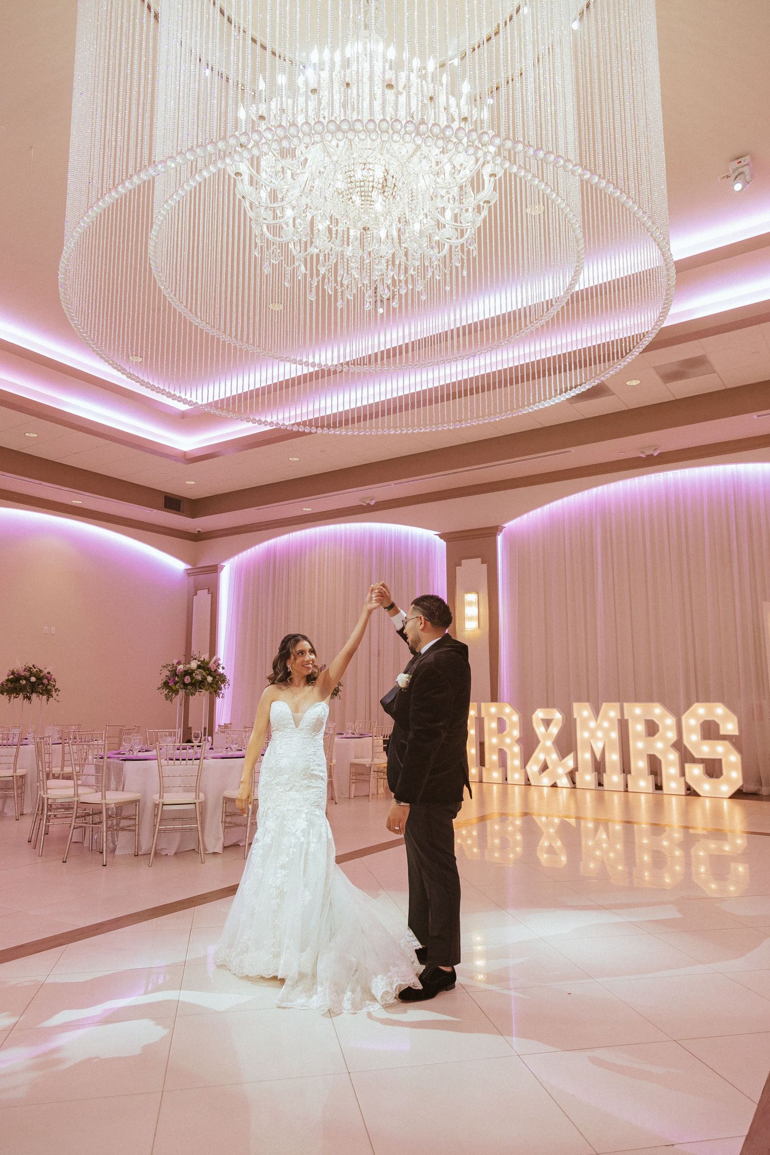 Wedding dance captured in luxurious ballroom with pink lighting and crystal chandelier overhead.