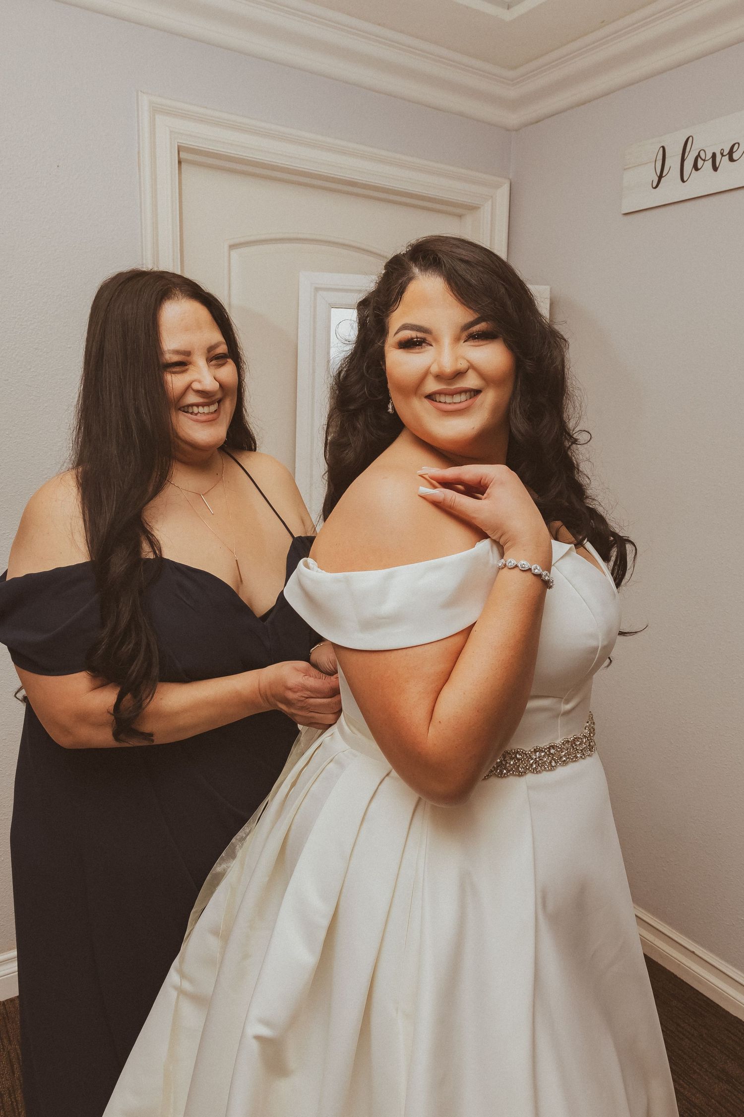 A joyful getting-ready moment between two women, one in a wedding dress and one in black.