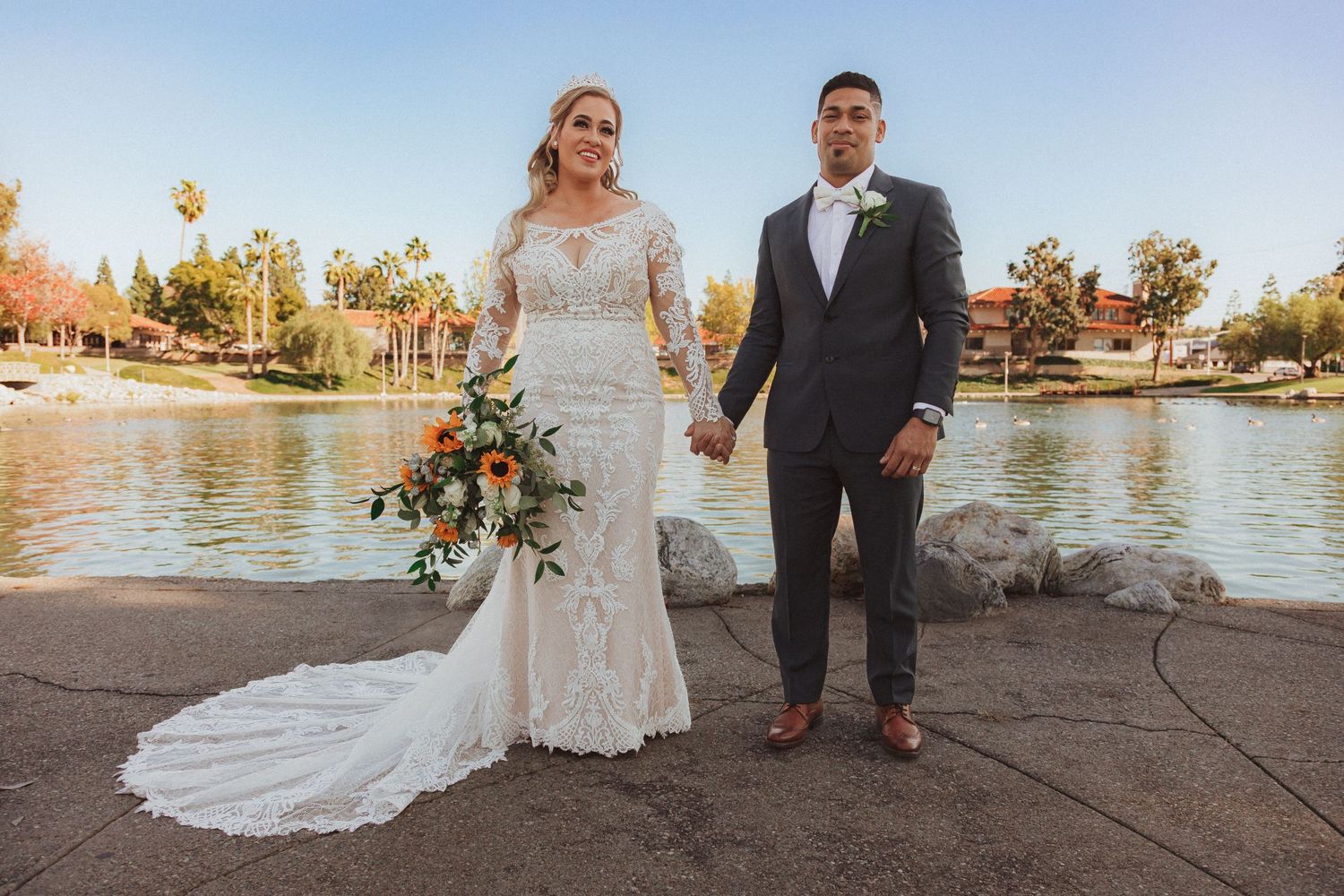 Wedding couple poses at sunset by a lake with autumn trees in background.