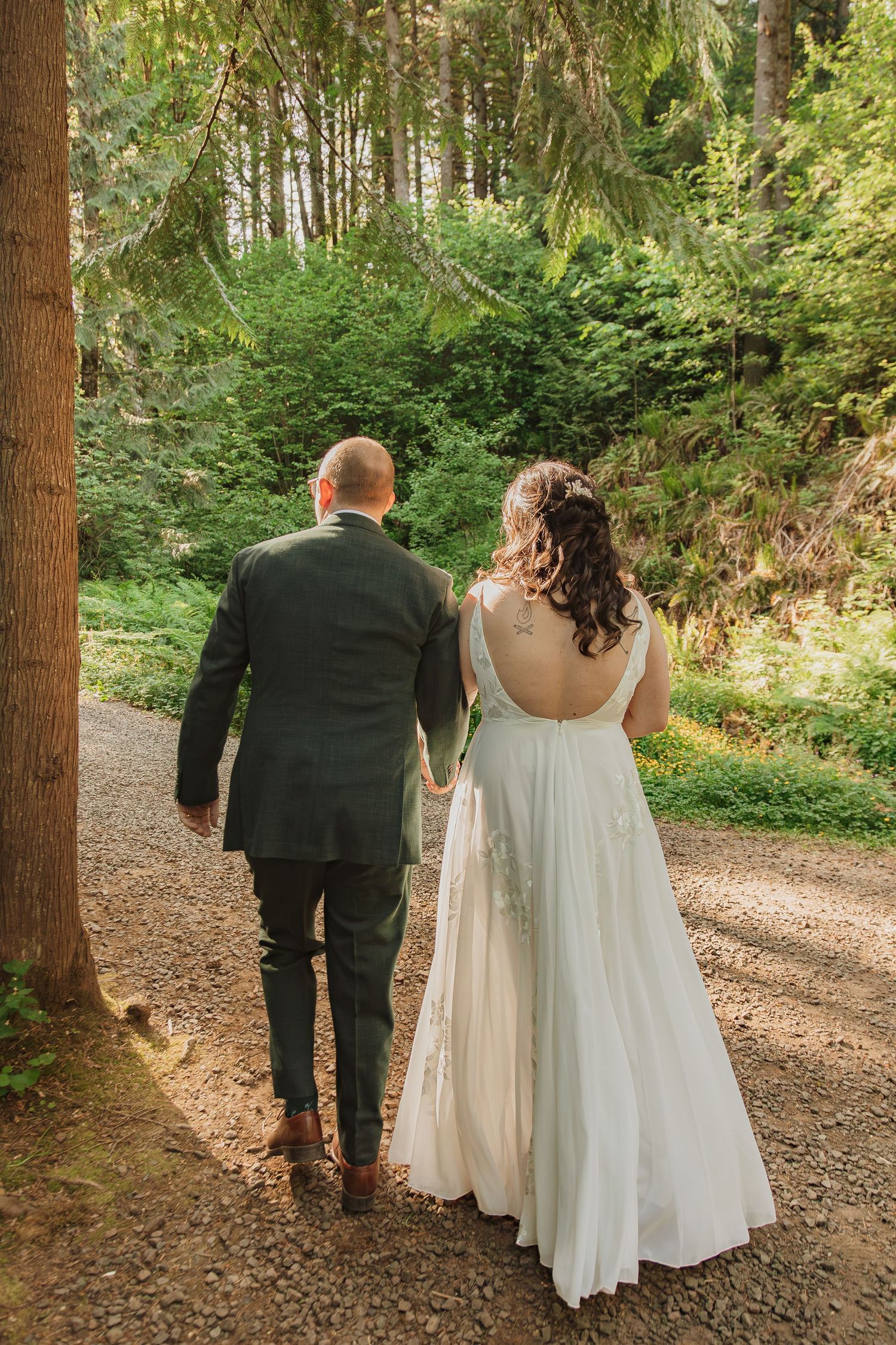 Bride in backless gown and groom walking together on a forest trail.
