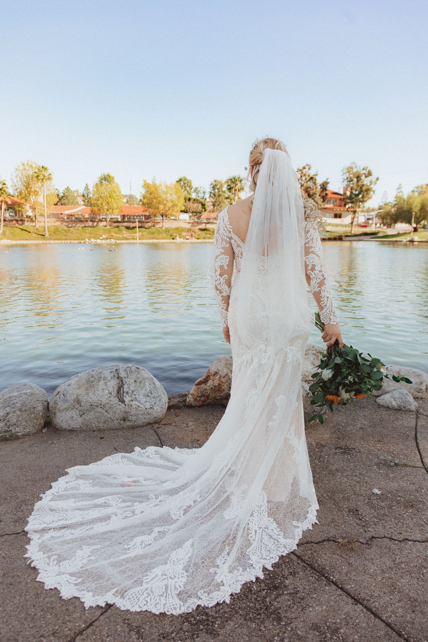 Long cathedral train and veil of lace wedding dress photographed from behind near water.