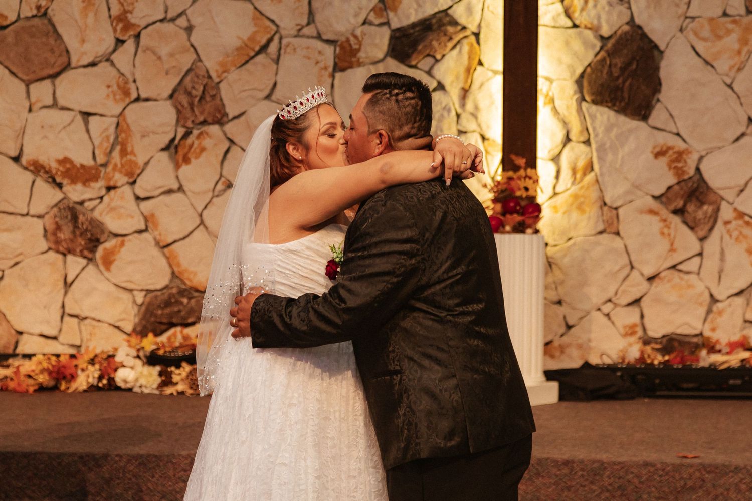 Newlyweds share their first kiss during a wedding ceremony in front of a stone wall backdrop.