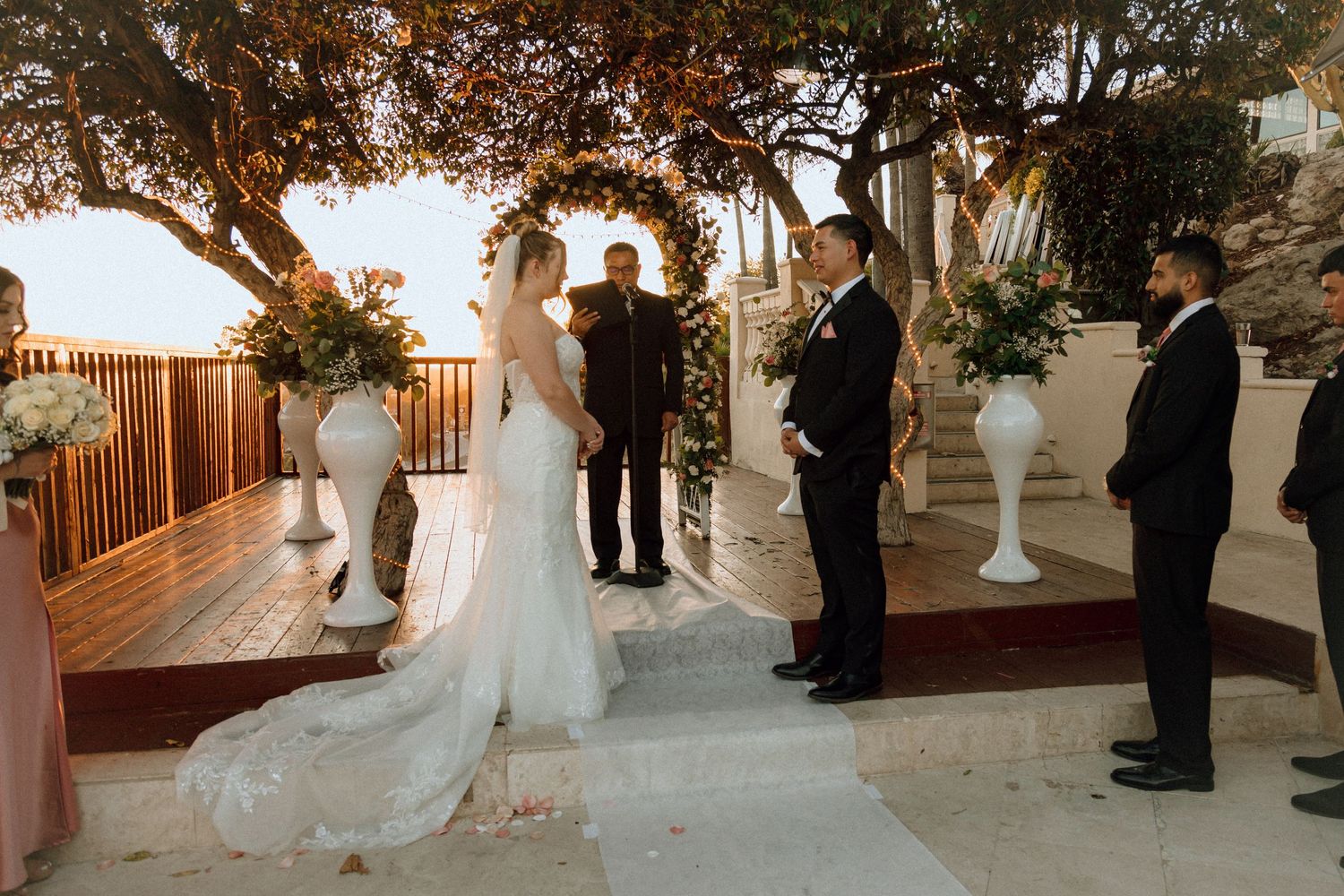 Wedding ceremony setup with wooden railing and white floral arrangements under natural tree canopy.