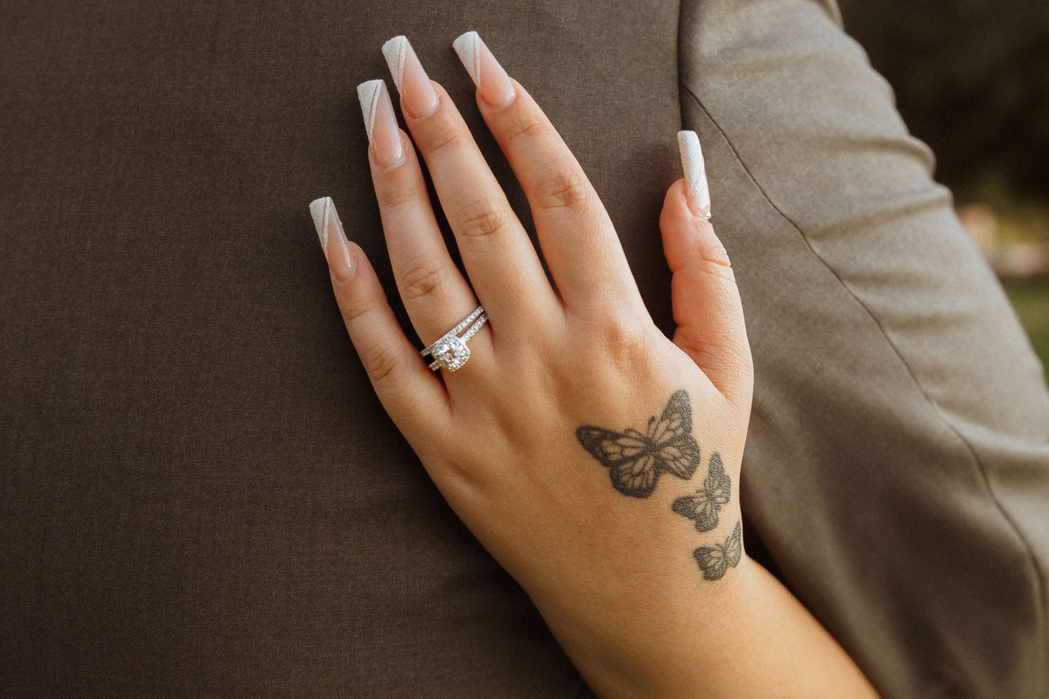 Manicured hand with butterfly tattoo and engagement ring against brown suit jacket.