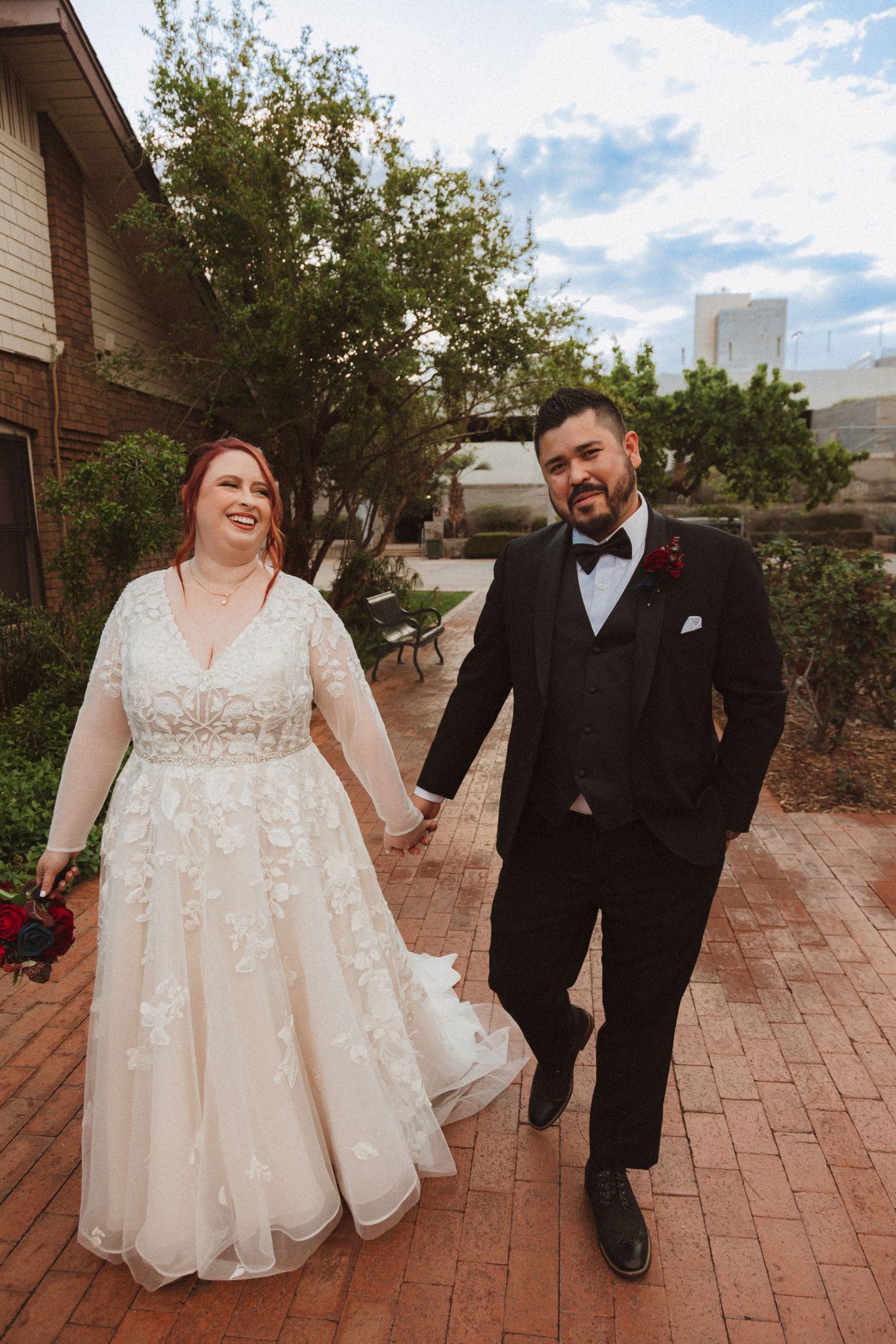 A joyful wedding couple shares a lighthearted moment on a brick walkway.