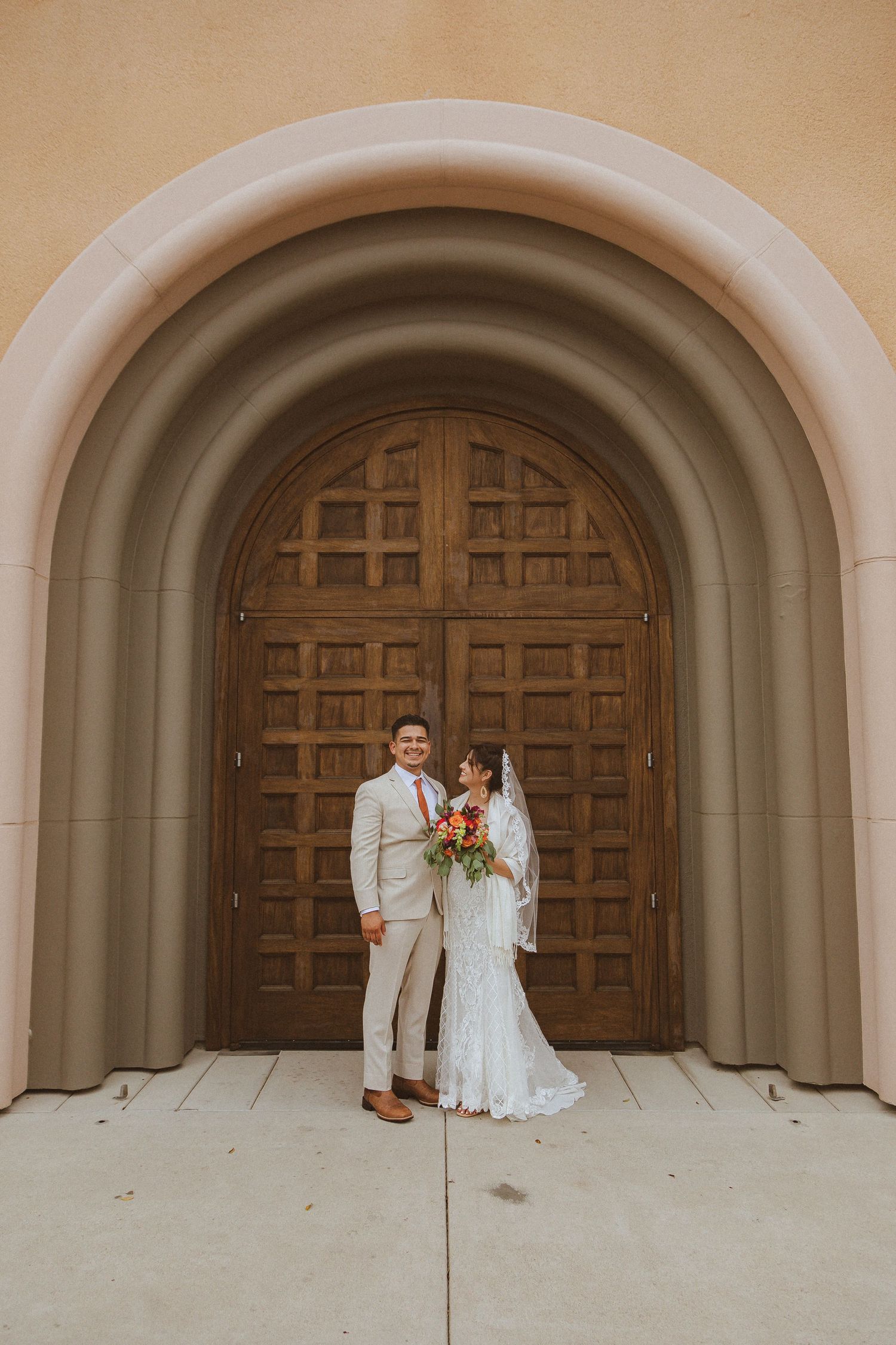 Wedding couple poses in front of grand wooden church doors under arched entryway.