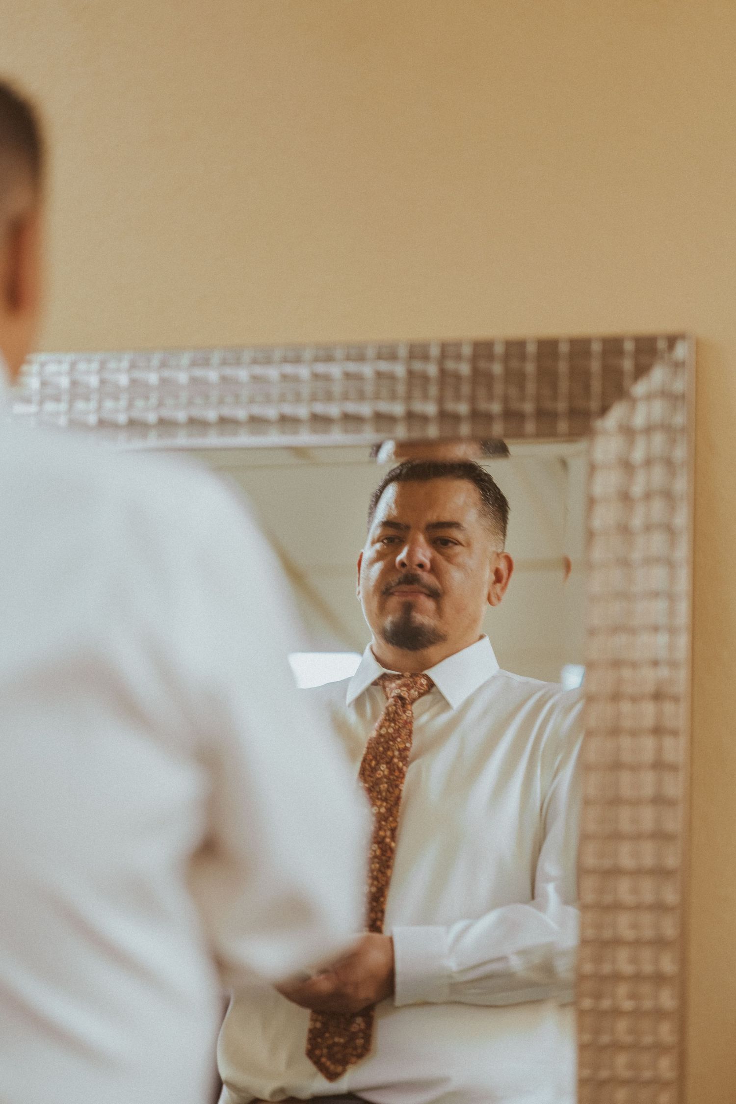 Person in white dress shirt and patterned tie looks in textured mirror while getting ready.