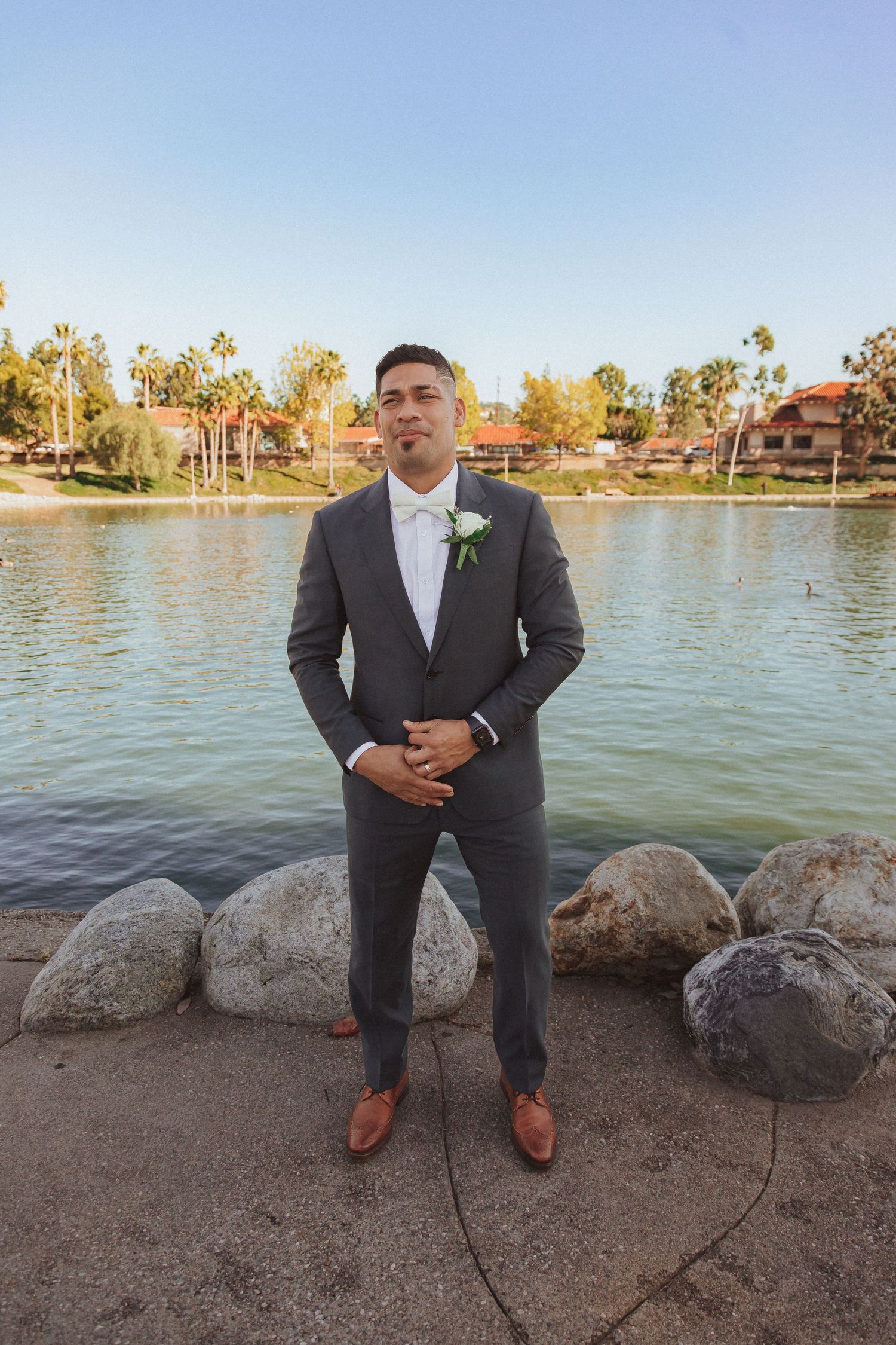 Portrait of groom in classic grey suit standing on stone pathway by lake.